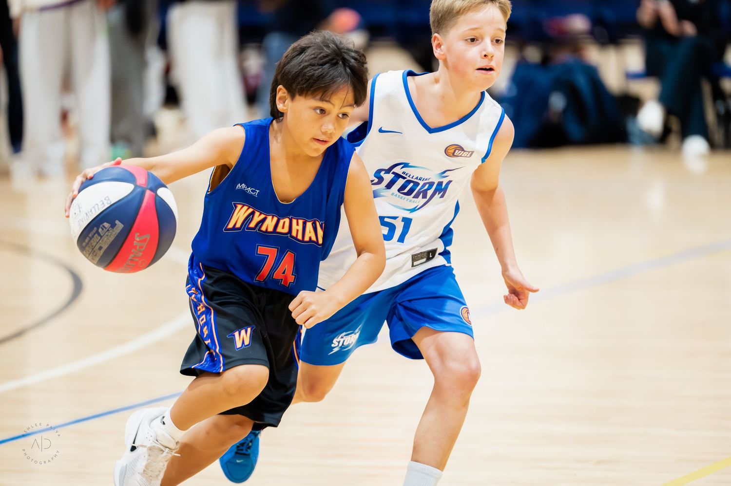 Two young boys play competitive basketball, one in a blue Wyndham jersey dribbling past a Storm player wearing number 51.