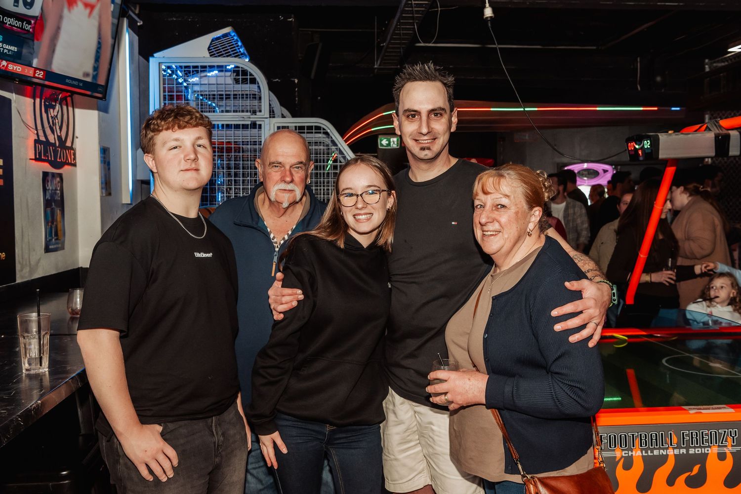 Group of five people posing together smiling in a bar arcade venue with colorful neon lights in background.