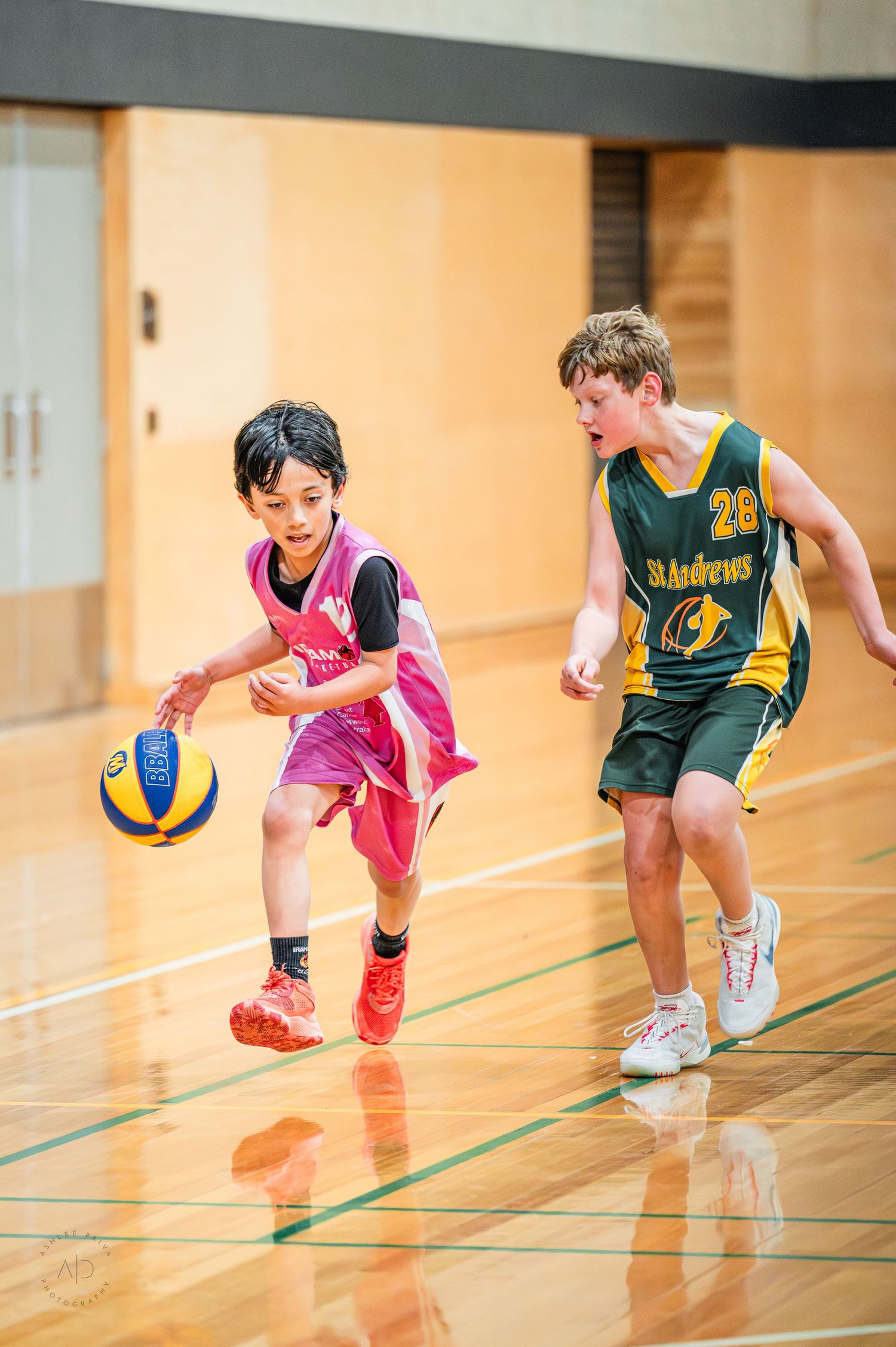 Two youth basketball players compete on an indoor court, one in pink dribbling past a St Andrews player in green.