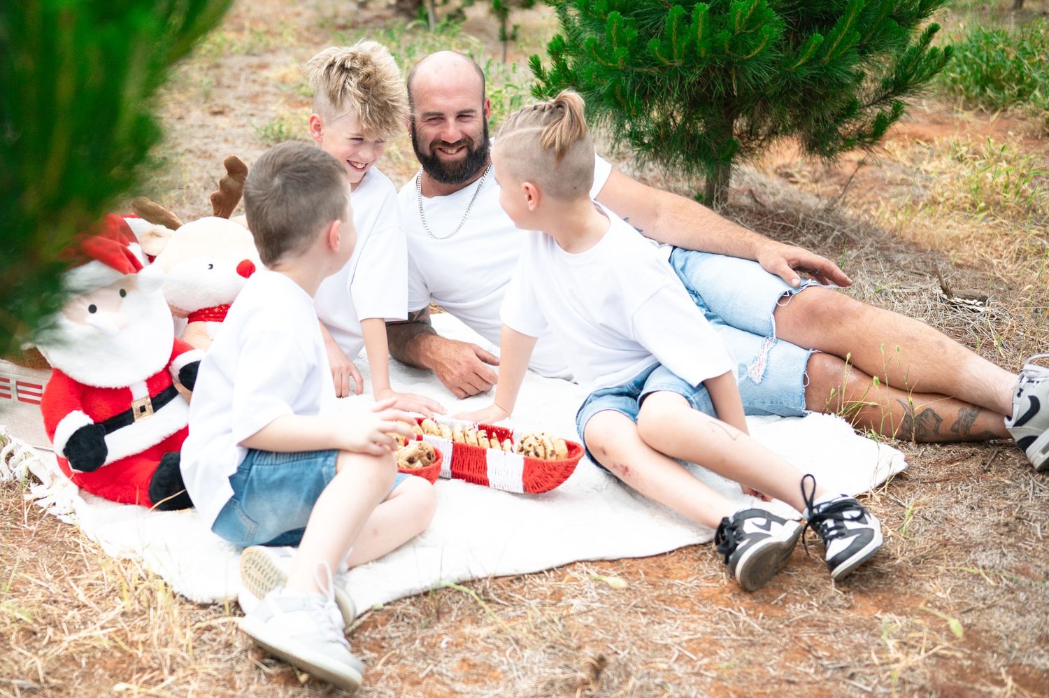 Family relaxing on a blanket outdoors near evergreen trees.