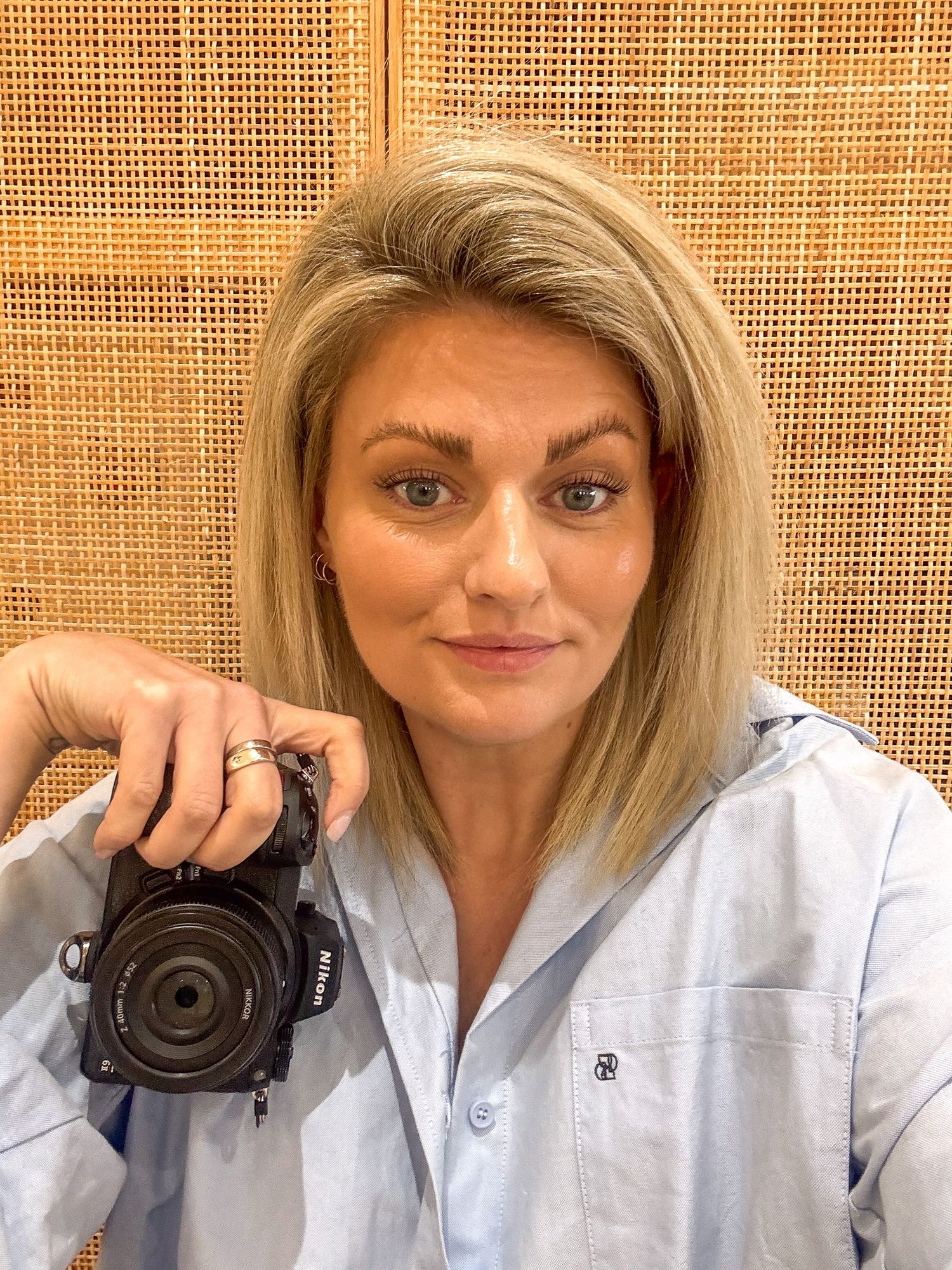 Blonde woman holding a Nikon DSLR camera, wearing a light blue shirt, posing in front of a rattan wall.