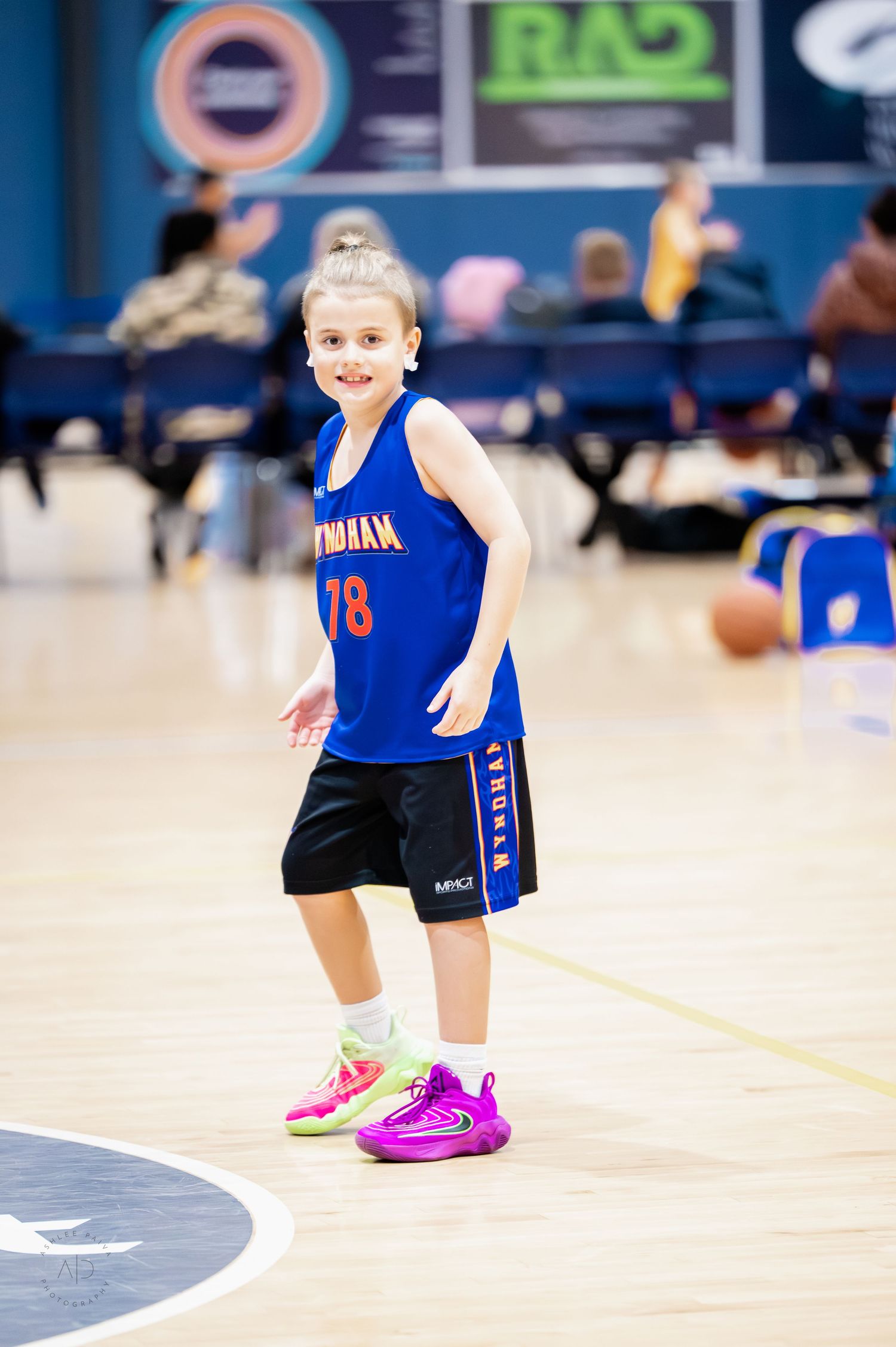 Young girl in blue number 78 basketball jersey smiles while playing on indoor court in colorful sneakers.