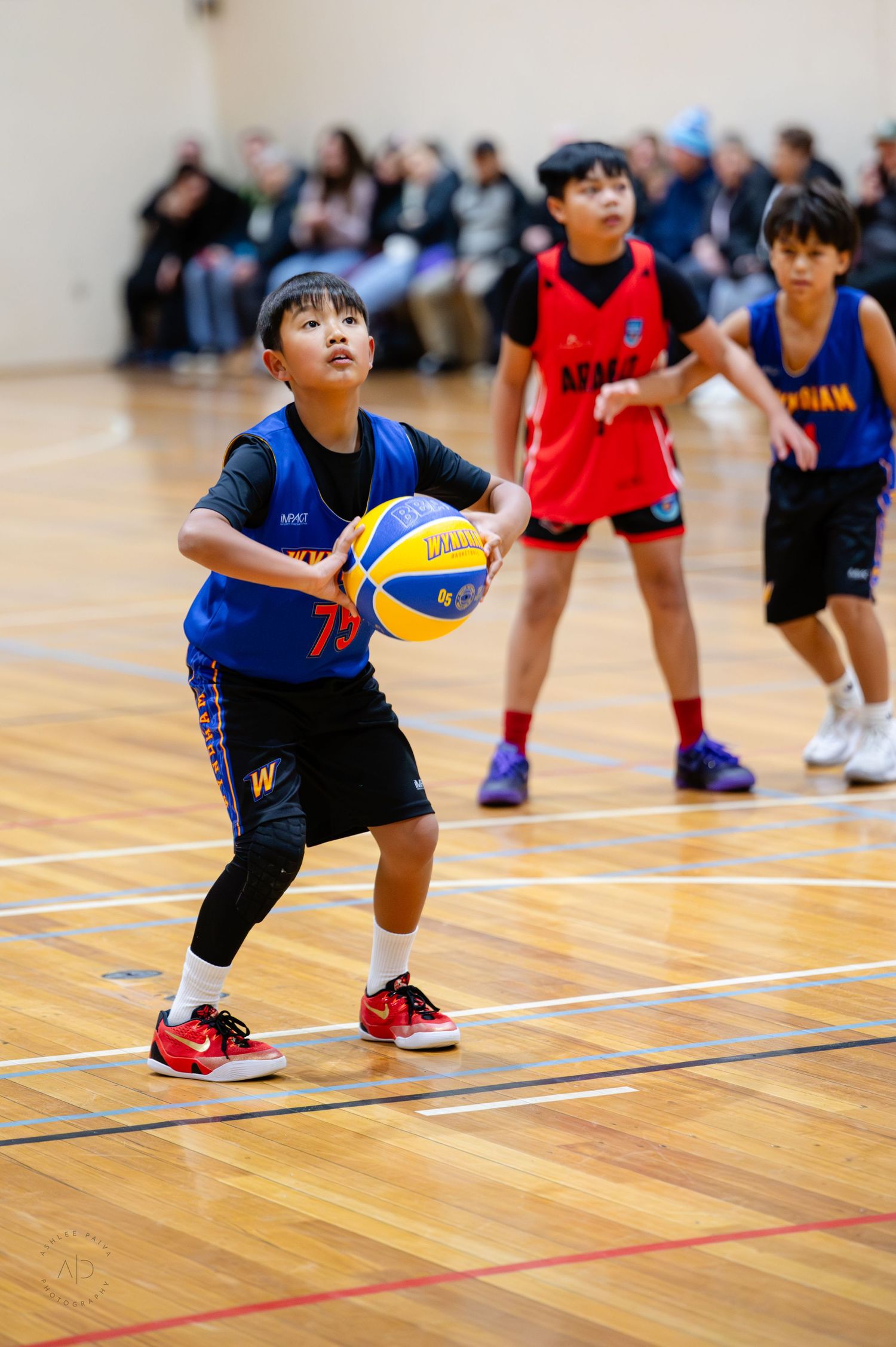 Young boy in blue jersey holds basketball during youth indoor basketball game with teammates in red uniforms.