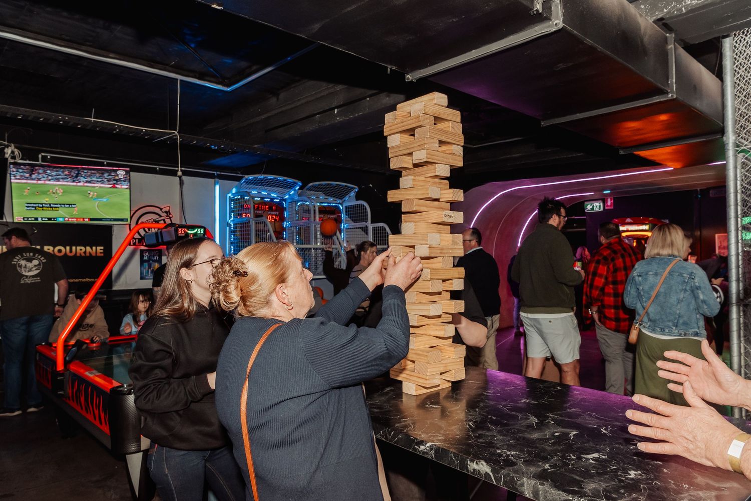 People playing giant Jenga at an arcade bar with colorful neon lights and arcade games in the background.