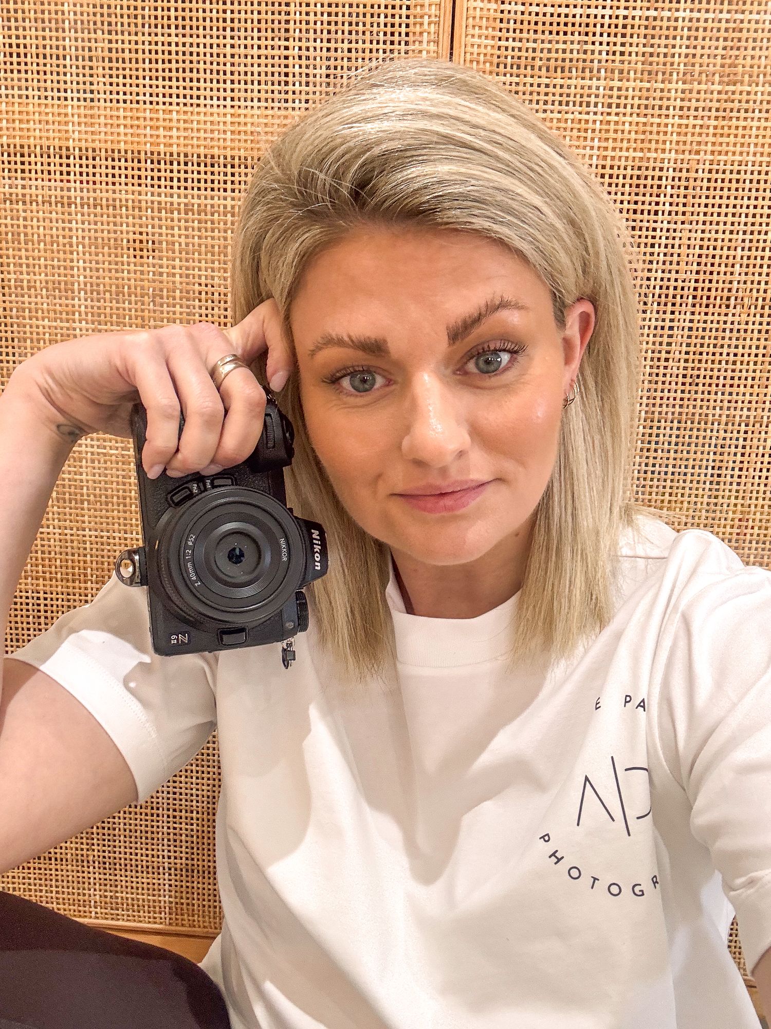 Blonde woman holding a Nikon camera up, wearing a white photography branded t-shirt, taking a selfie indoors.
