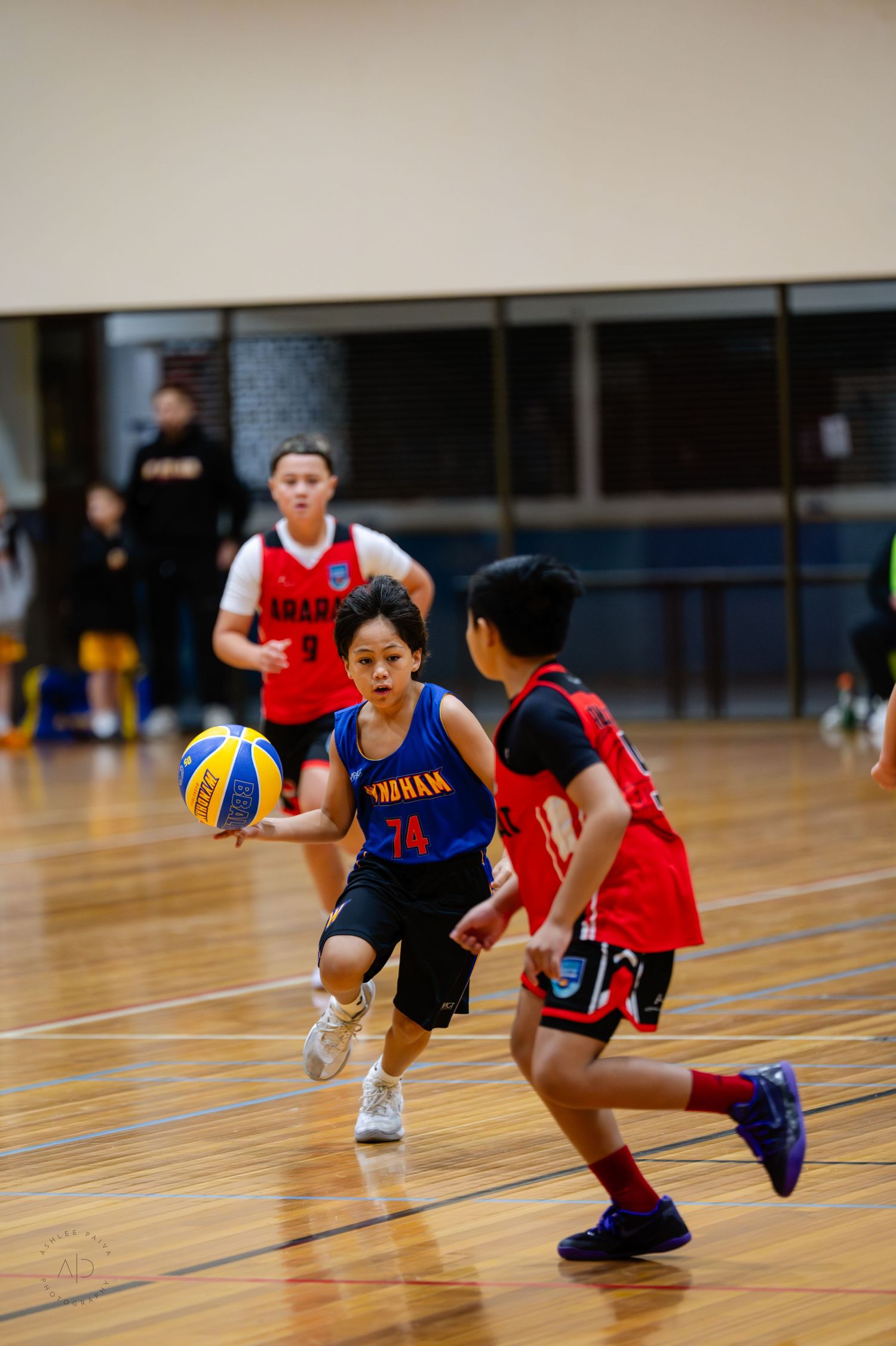 Young basketball players compete in an indoor court game, with player #74 in blue dribbling past defenders.
