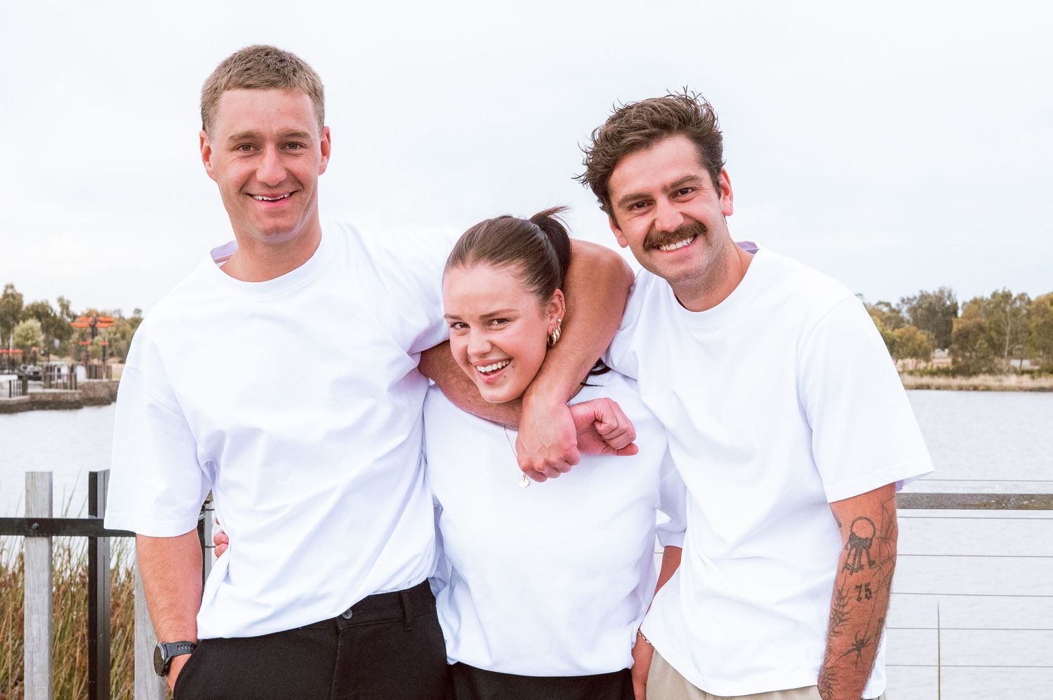 Three people in white shirts share a playful moment together on a waterfront deck.