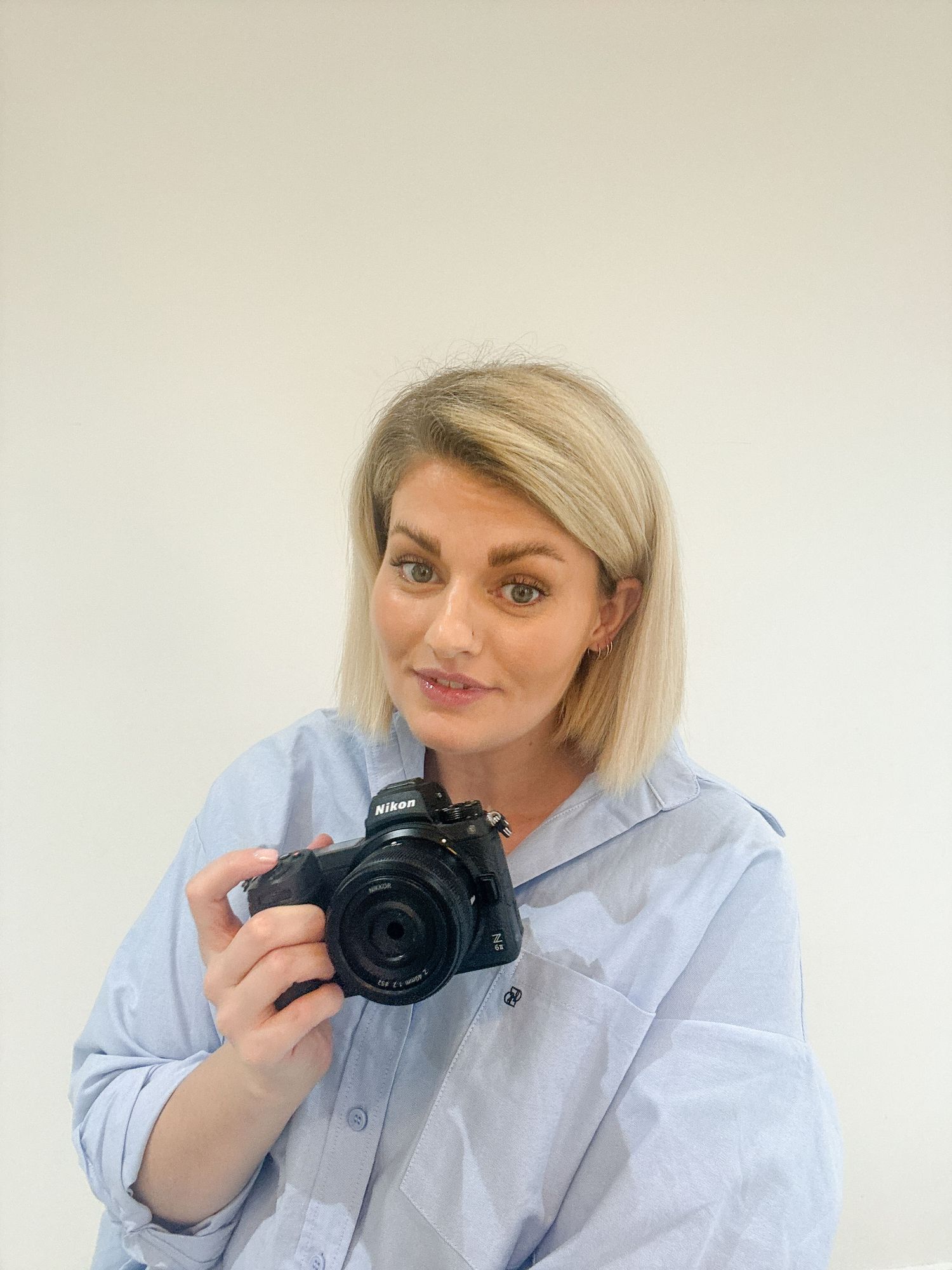 Photographer in light blue shirt posing with professional camera against white wall.