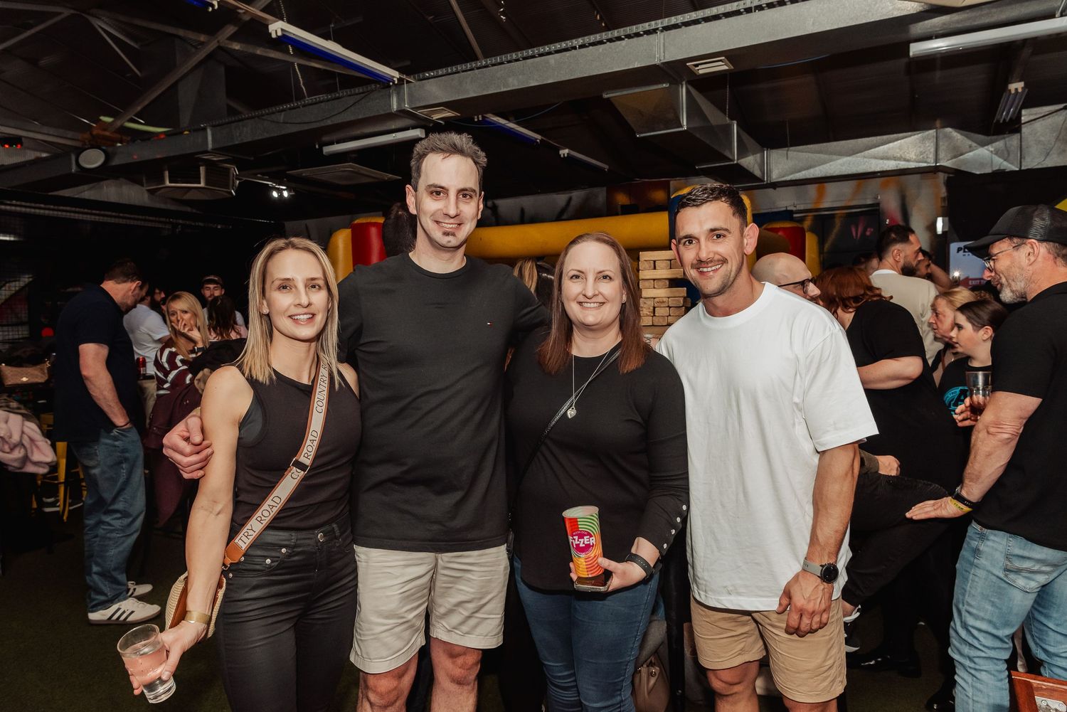 Four friends smiling together at a lively indoor event, dressed casually in black tops and khaki shorts.