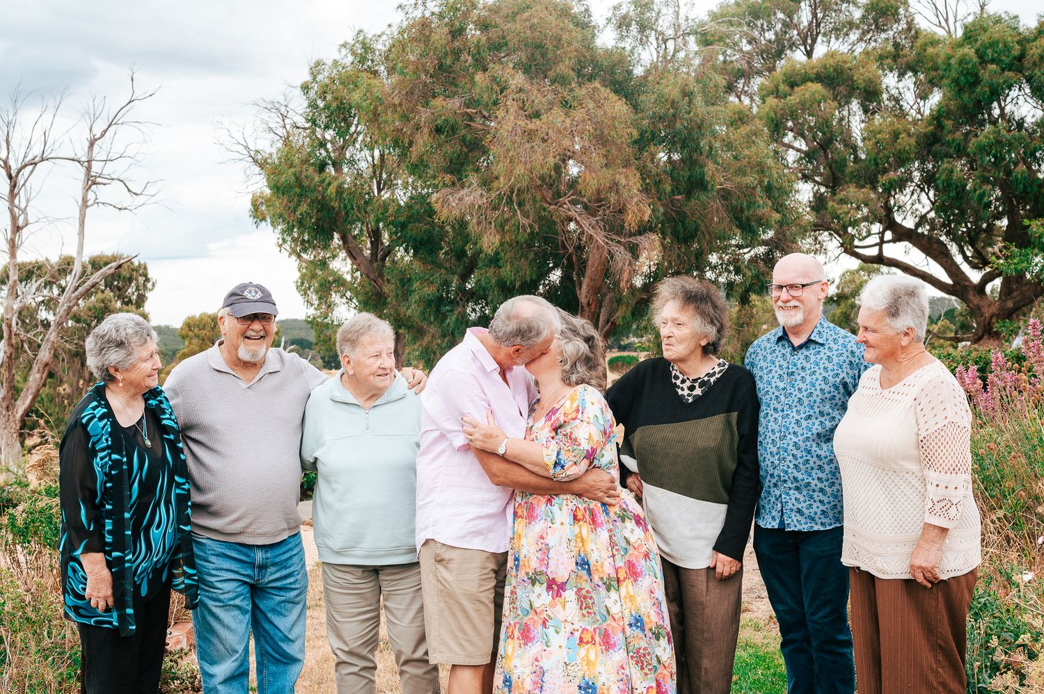 A group of senior friends gather outdoors in a garden setting on a cloudy day.