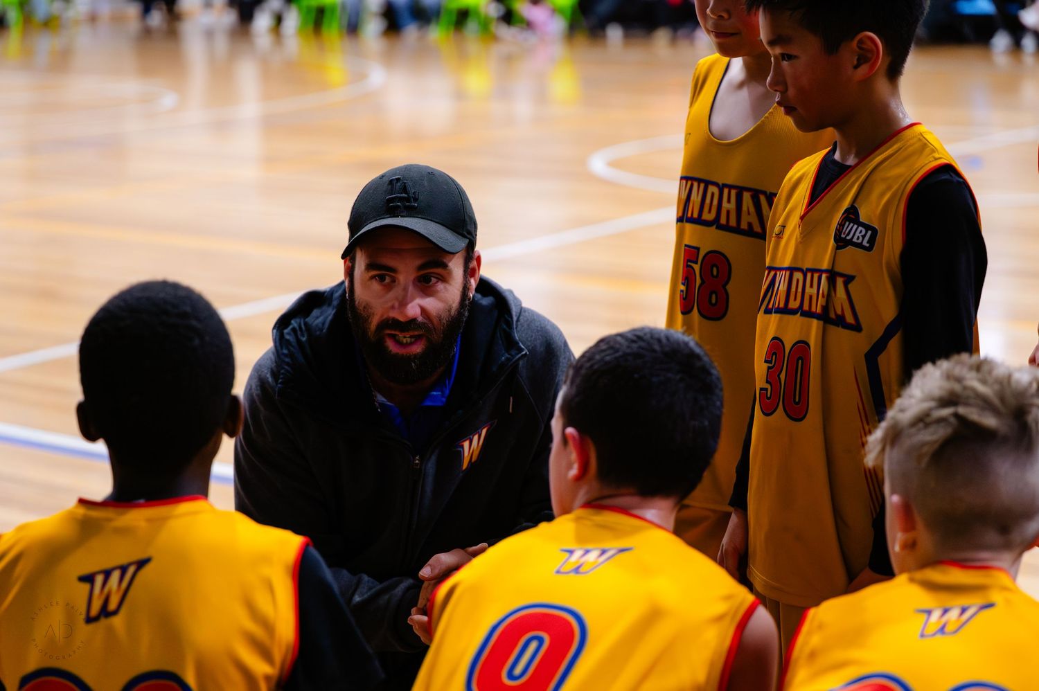 Basketball coach giving a timeout huddle talk to young players in yellow jerseys on an indoor court.