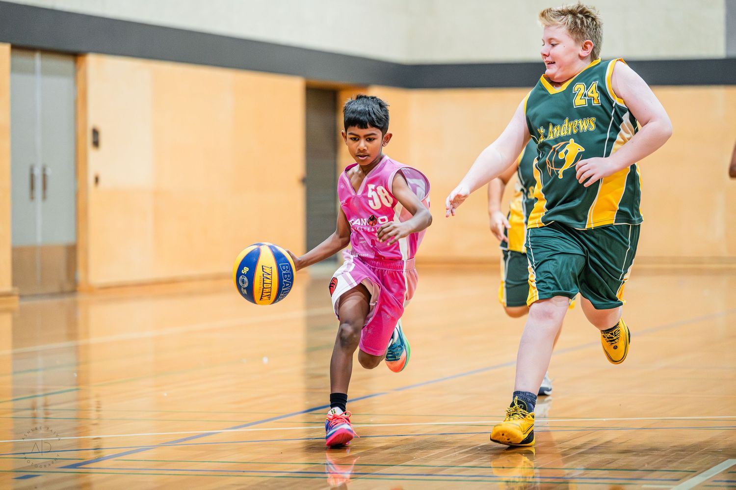 Two young children playing basketball indoors, one in pink dribbling past a defender in green uniform.