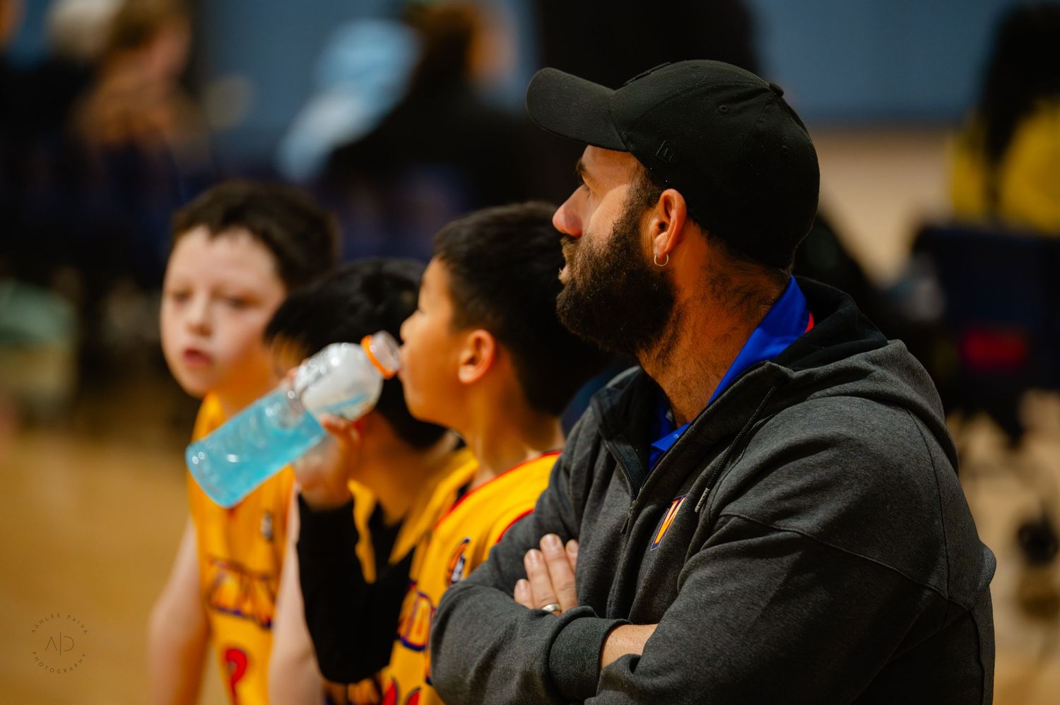 Adult male coach with beard and cap watches youth basketball game intently, with young players in yellow jerseys nearby.