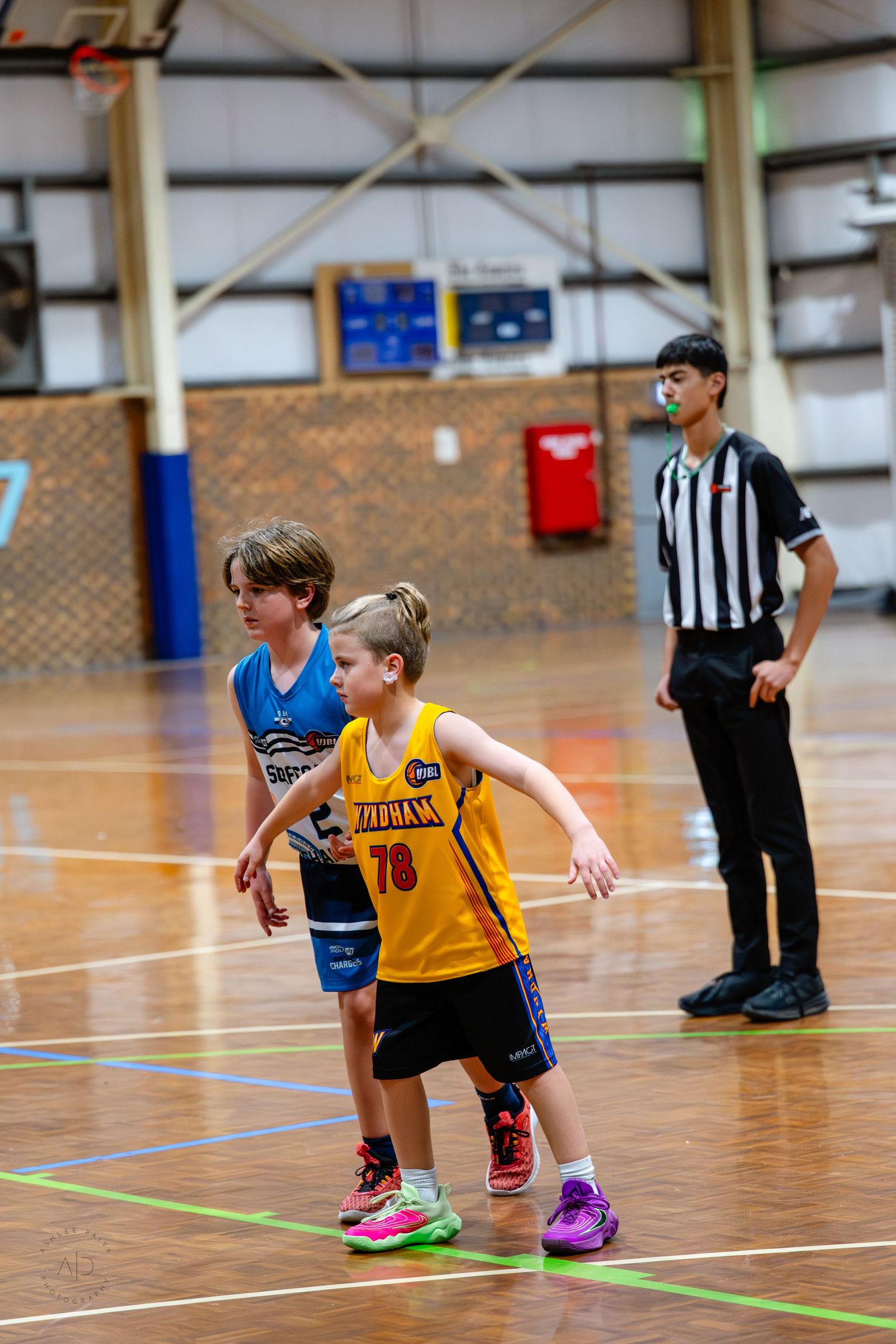 Two young boys play basketball in yellow and blue jerseys as a teenage referee watches during an indoor youth league game.