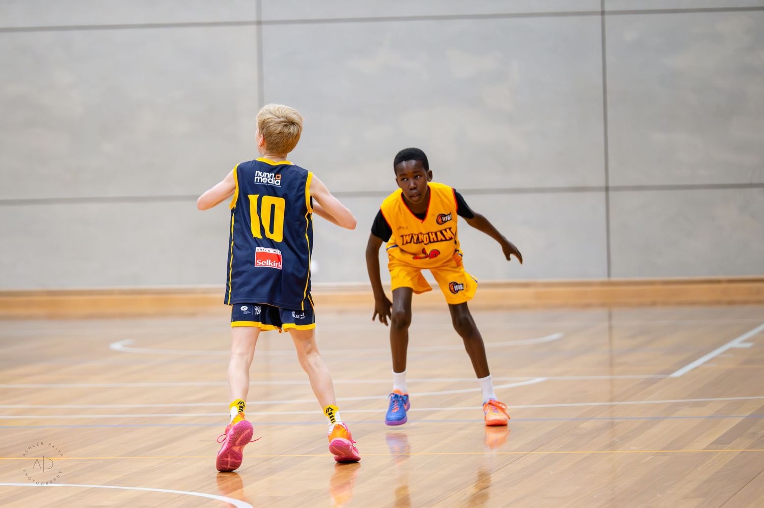 Two youth basketball players compete on an indoor court, one in blue jersey number 10, the other in yellow uniform.