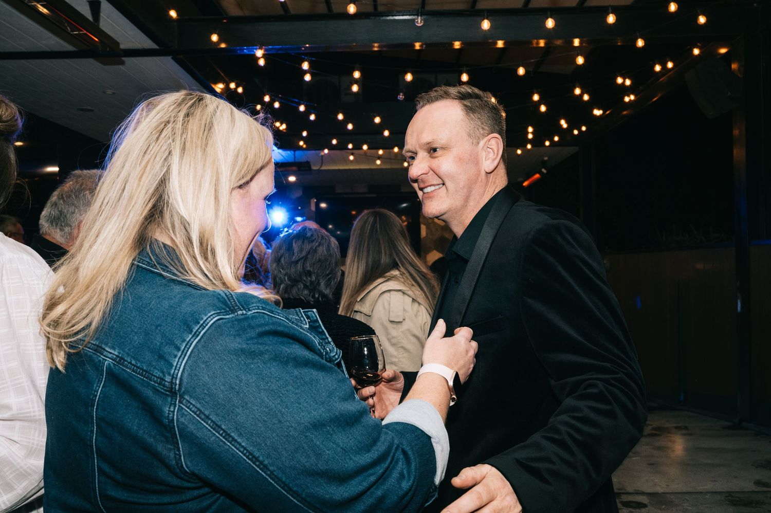 Party guests dancing and socializing at evening celebration under string lights.