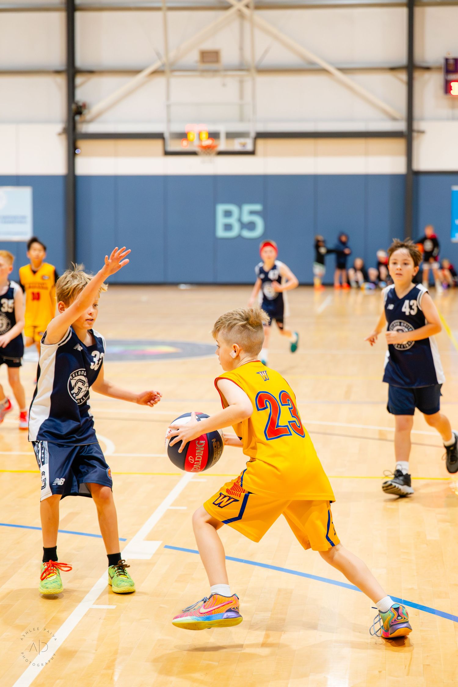 Young boys playing youth basketball in an indoor court, with player #23 in yellow dribbling past a defender.