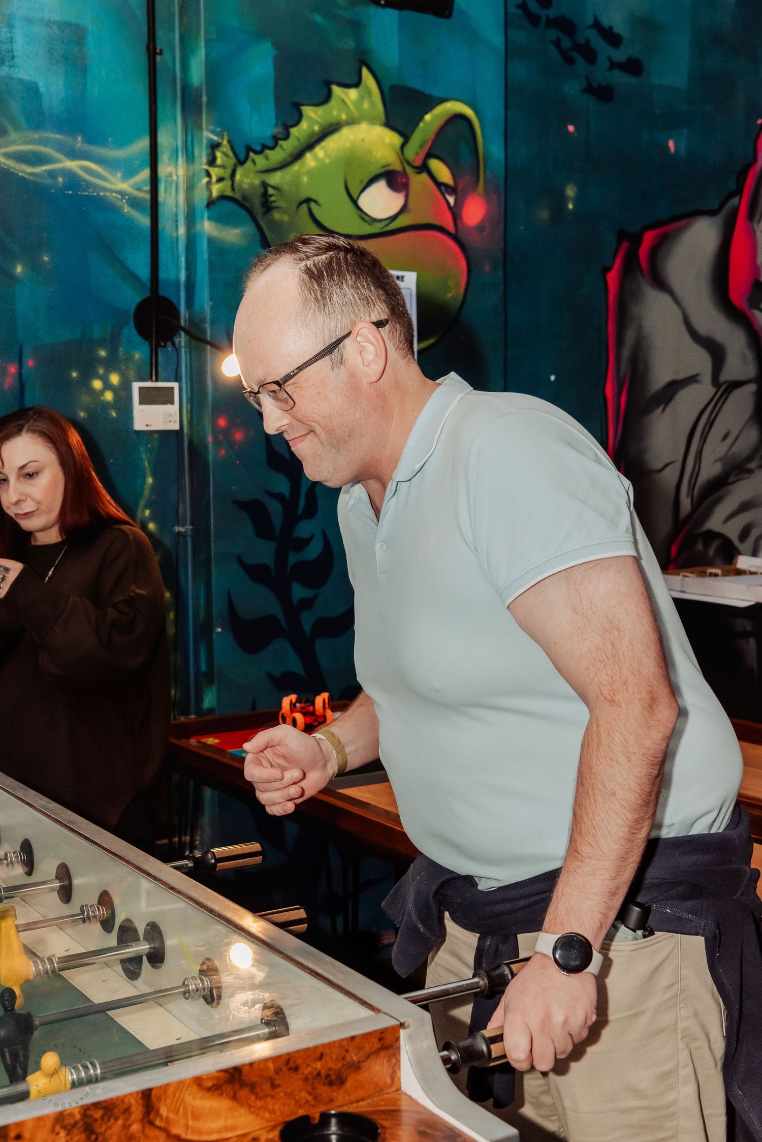 Man in light blue polo shirt playing foosball in an arcade bar with colorful graffiti mural background.