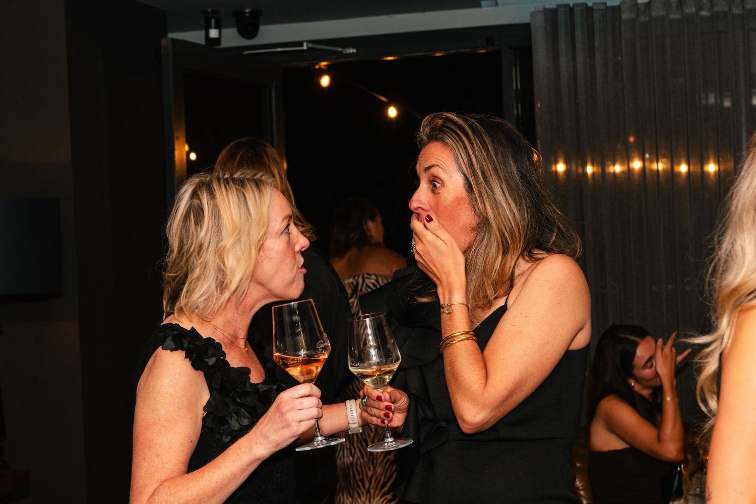 Two women holding wine glasses at a party, one covering her mouth in surprise during a lively conversation.