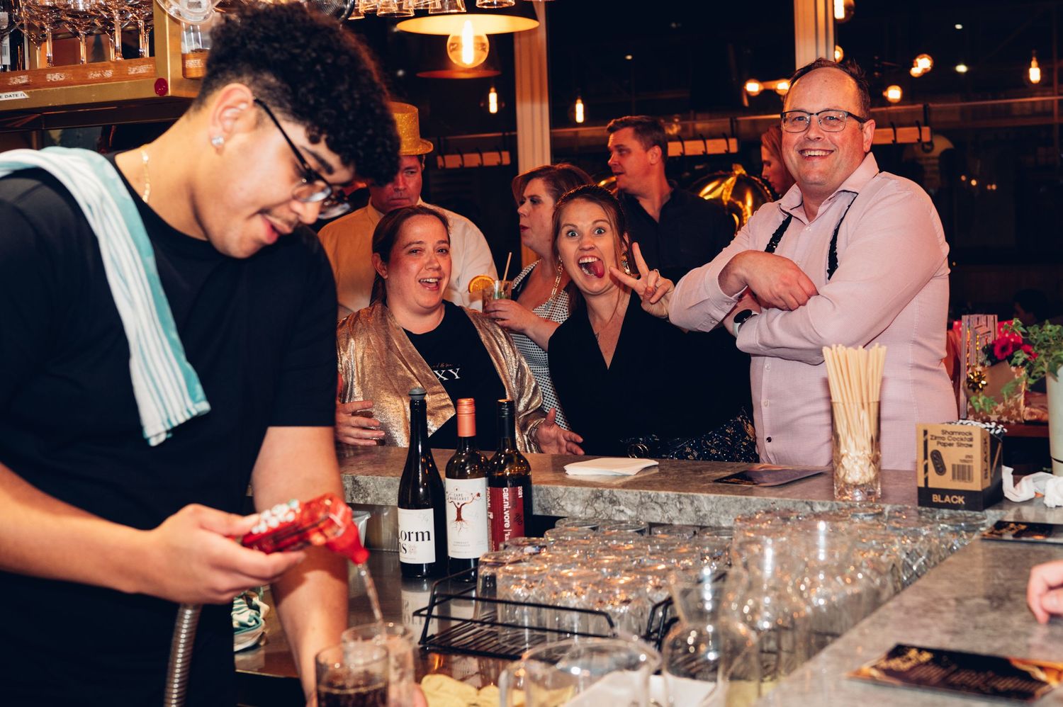 Bartender preparing cocktails while guests socialize at an illuminated bar counter.