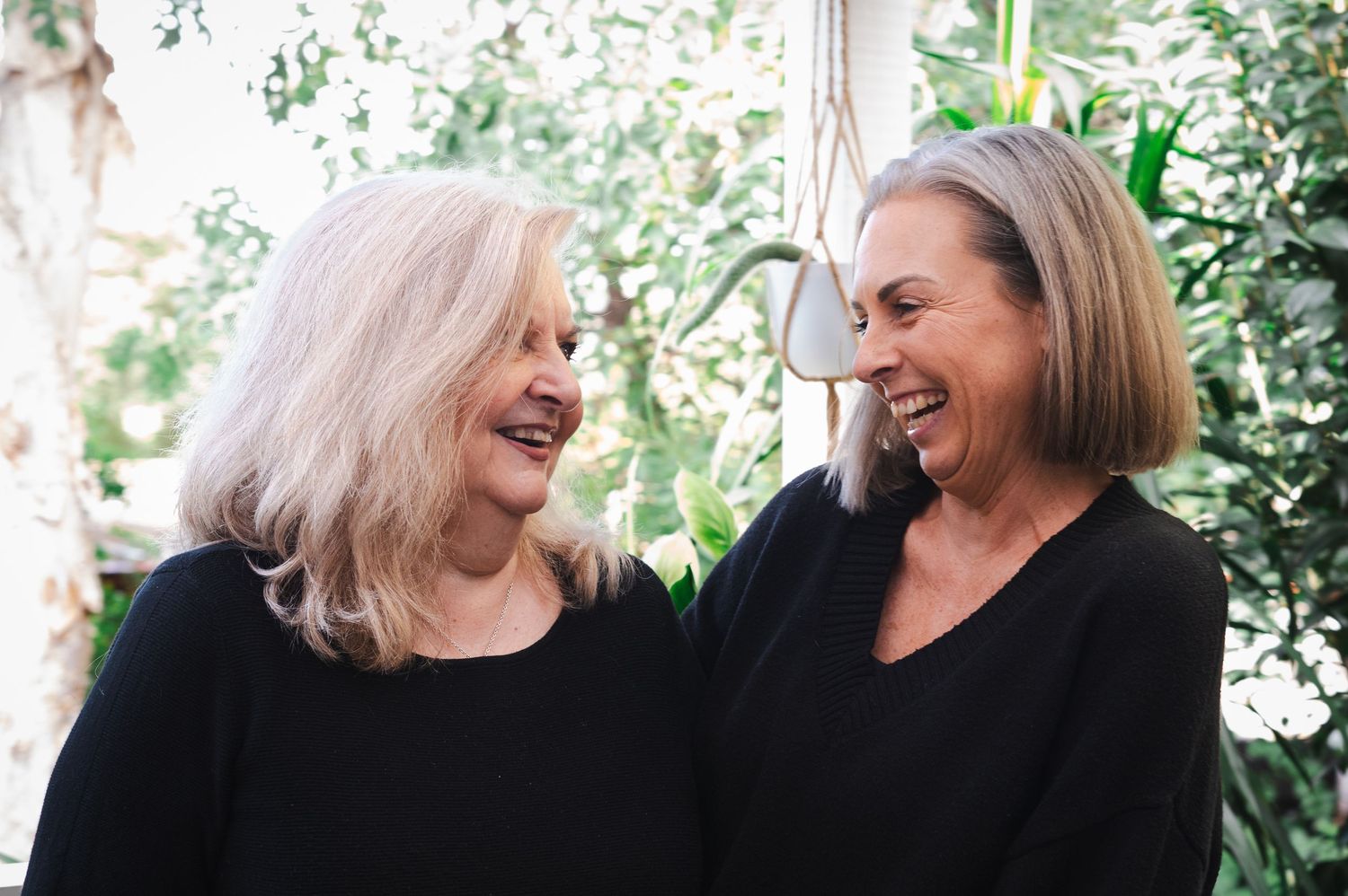 Two older women in black tops laughing together outdoors, surrounded by lush green plants.
