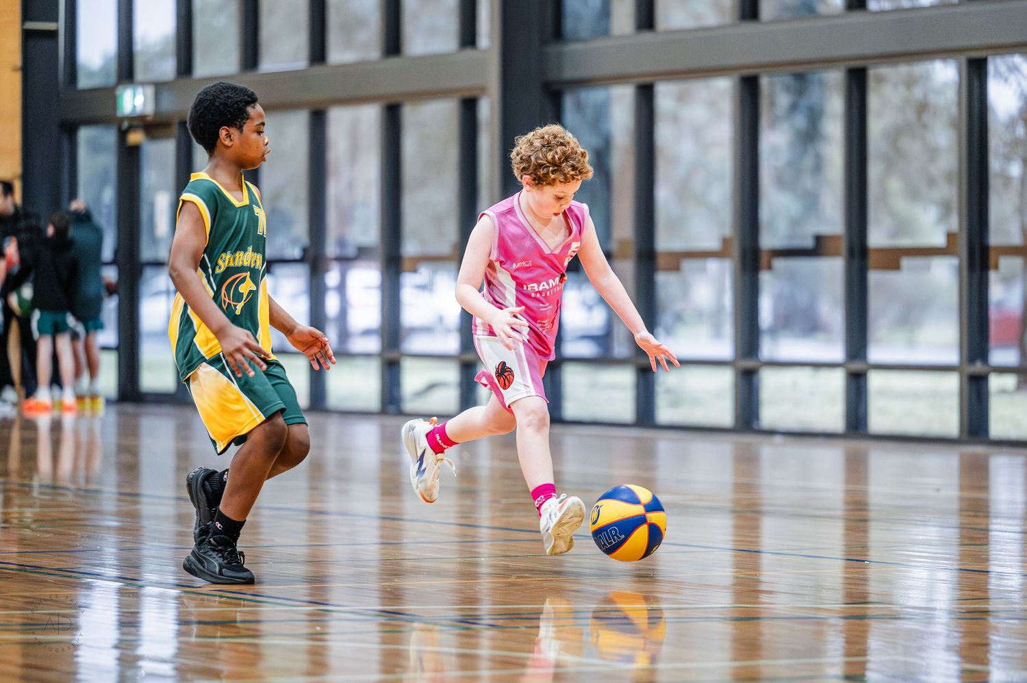 Two young boys playing basketball indoors, one dribbling in pink uniform, other defending in green and yellow.