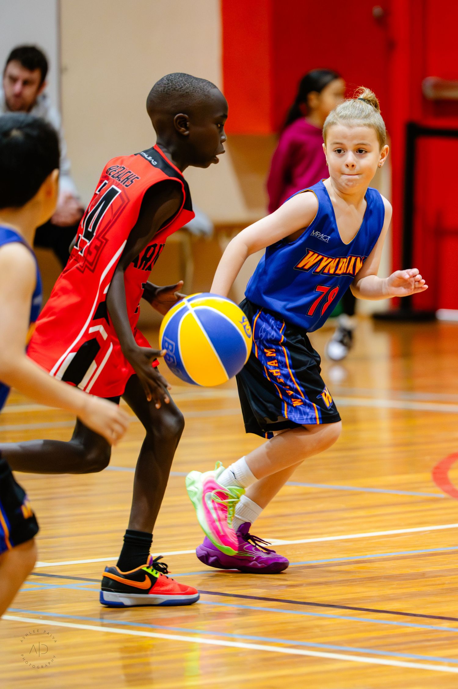 Two young basketball players compete on an indoor court, one dribbling in a blue Wynaham jersey, the other defending in red.
