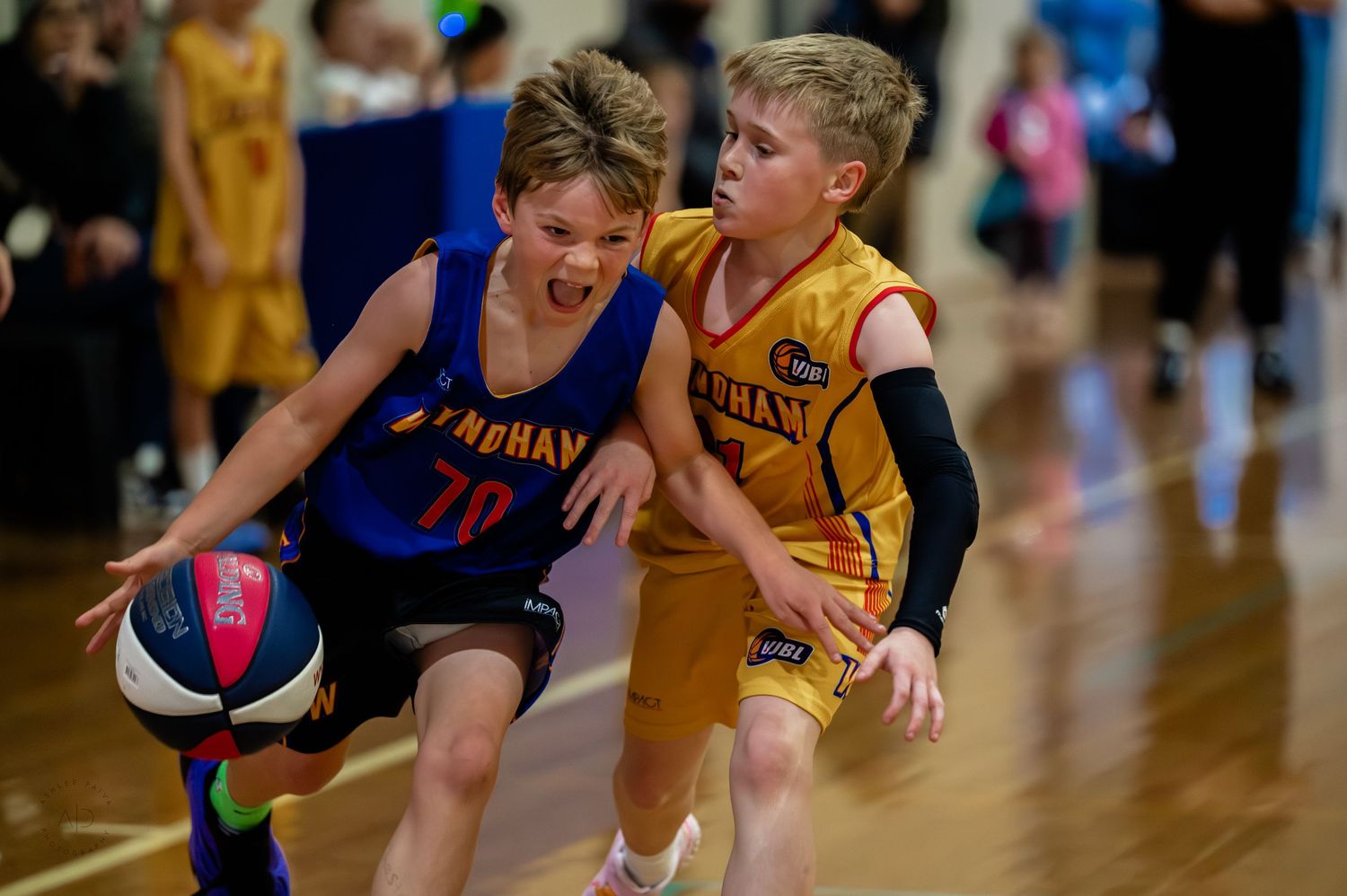 Two young boys playing competitive youth basketball, one in blue uniform dribbling past a defender in yellow.