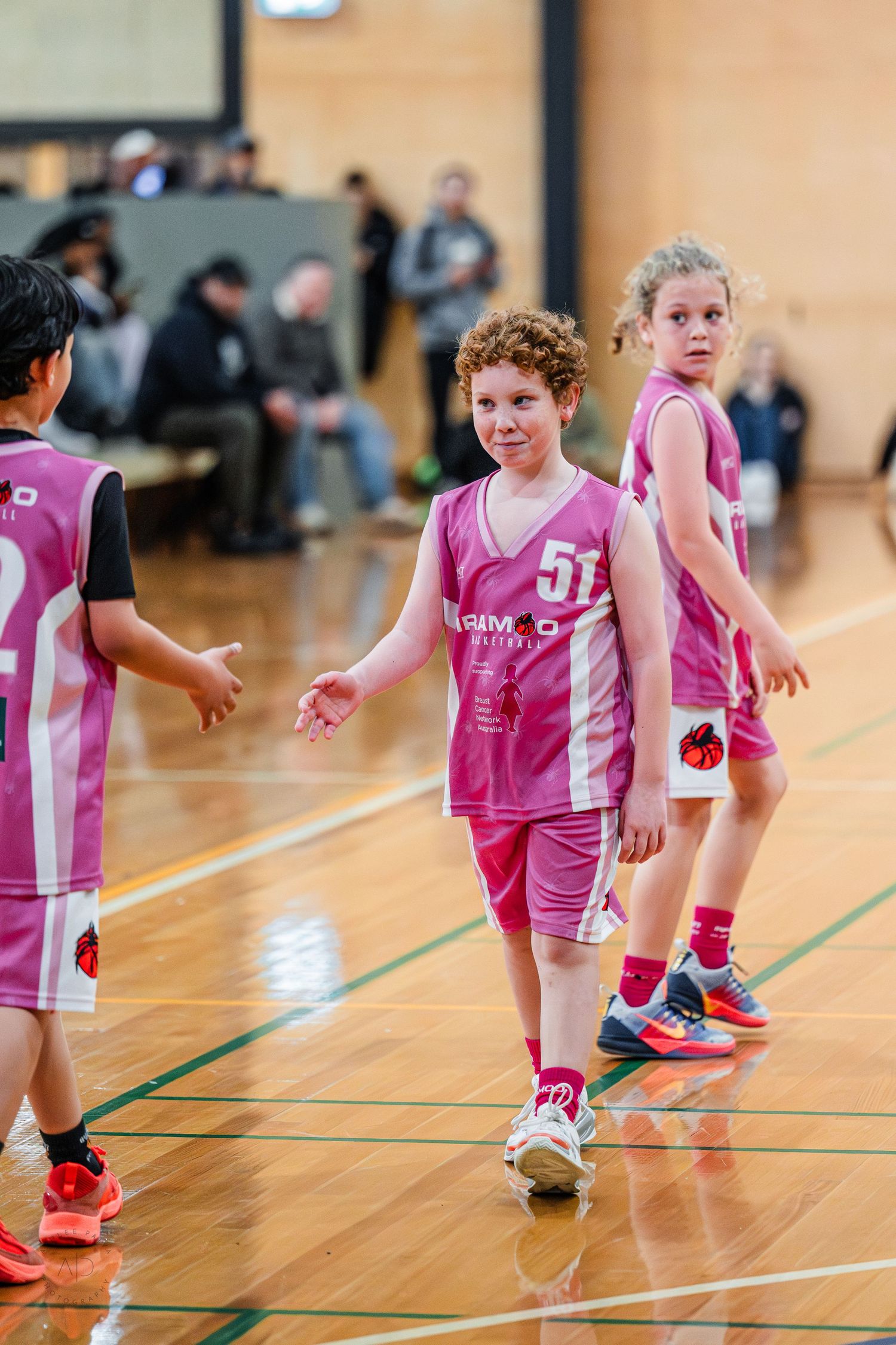 Young basketball players in pink uniforms compete in an indoor youth basketball game on a wooden court.