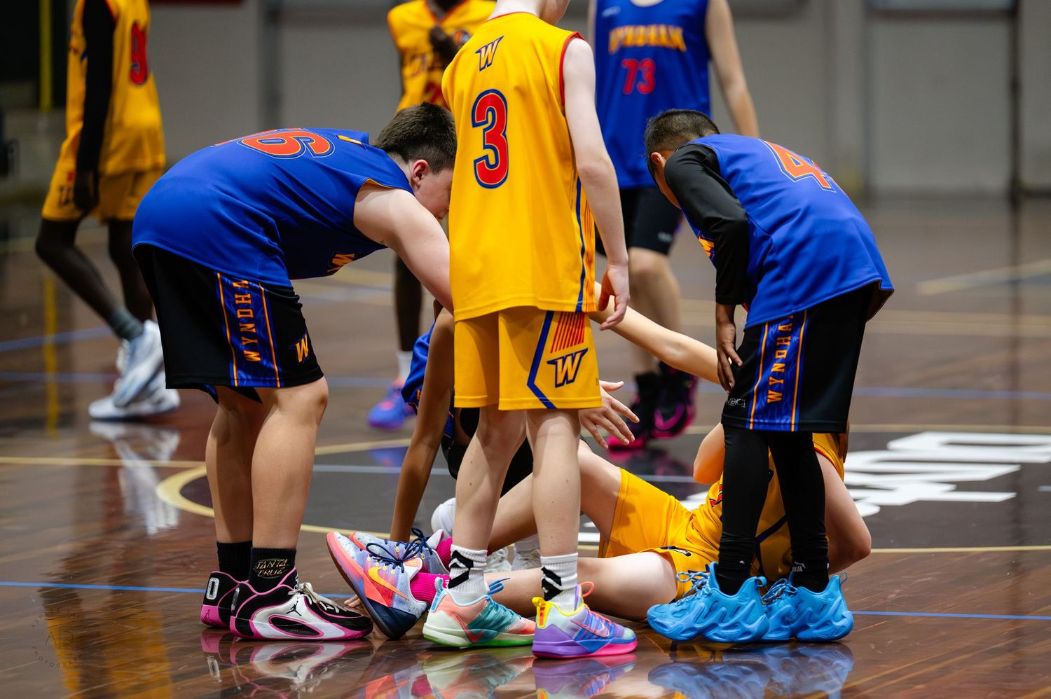 Basketball players in blue and yellow uniforms huddle around a fallen player on an indoor court during a youth game.