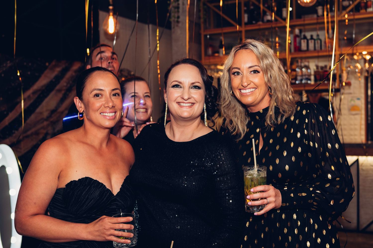 Three friends in formal attire pose together at an evening celebration with string lights.