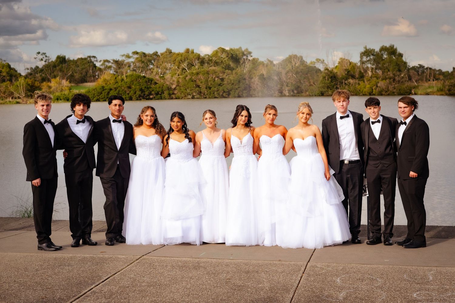 Group of teens in formal wear, girls in white gowns and boys in tuxedos, posing outdoors near a lake.