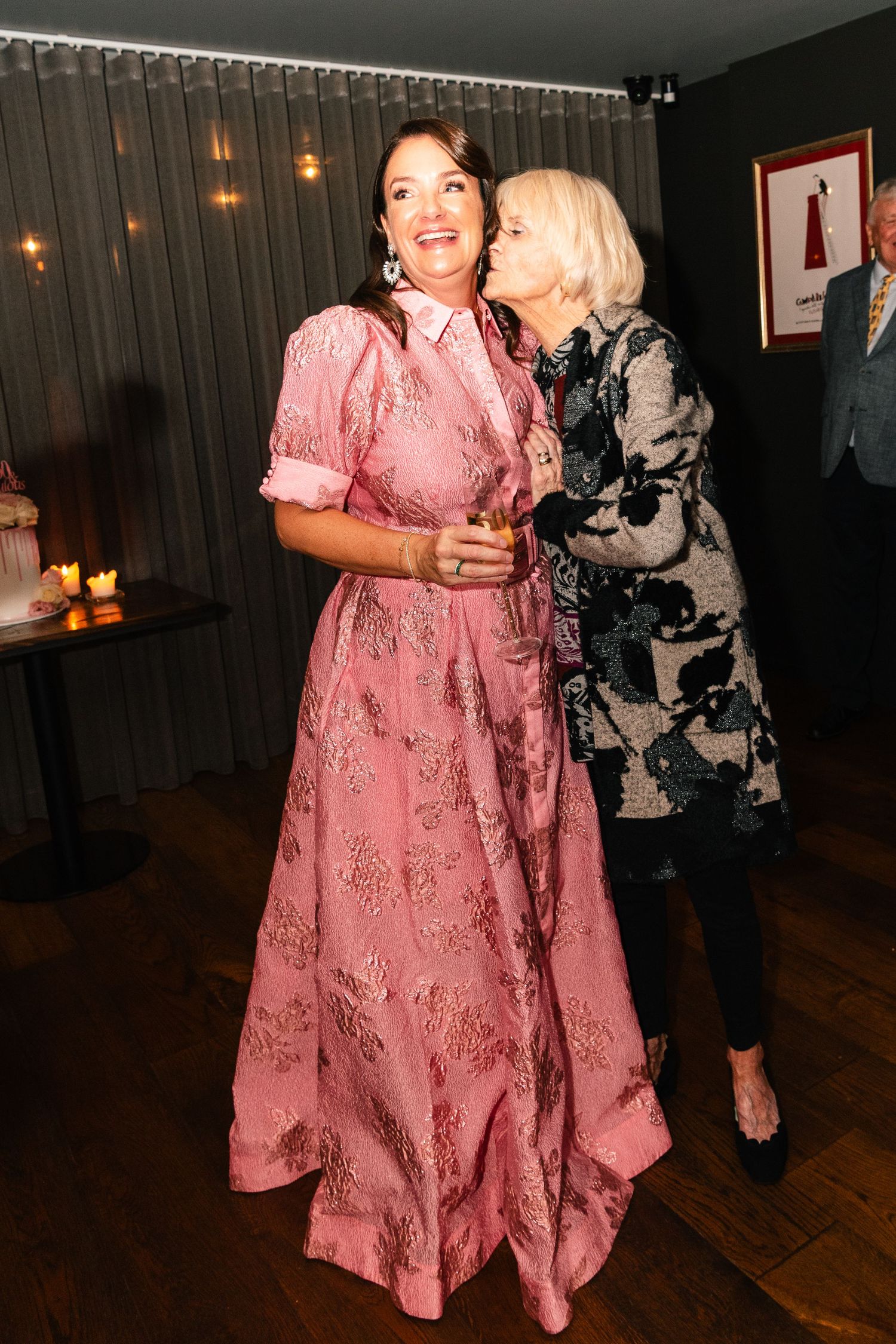 Two women at a party, one in a pink floral gown, the other in a floral jacket, sharing a warm moment.