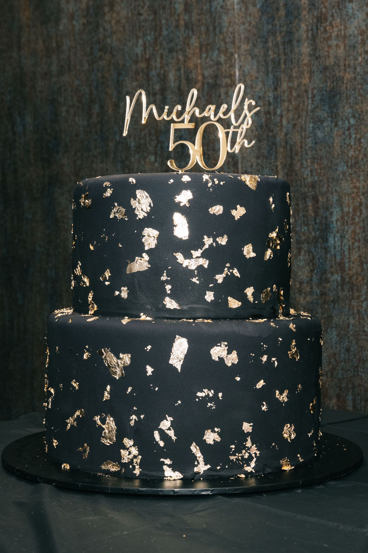 Elegant black and gold two-tier 50th birthday cake with metallic accents and custom topper.