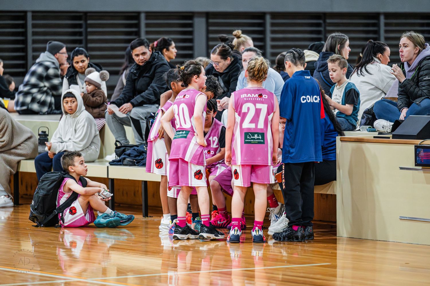 Women's basketball team in pink uniforms huddle with coach during timeout on indoor court.