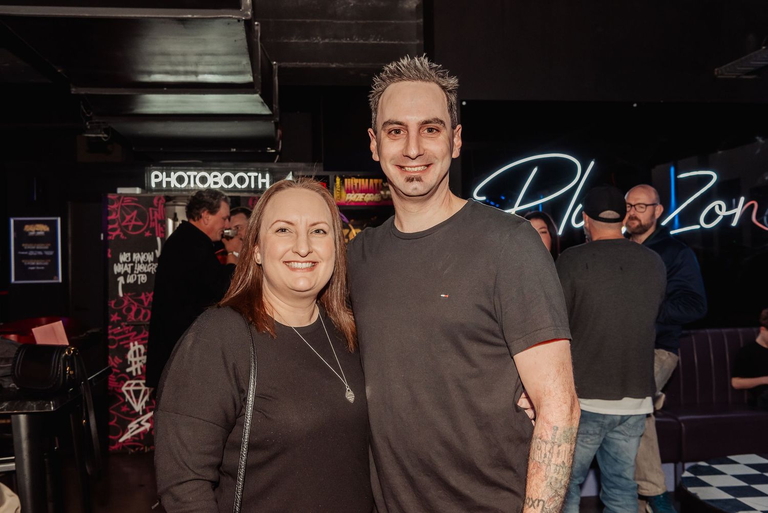 Two people smiling together in front of a photobooth sign at a social event venue.