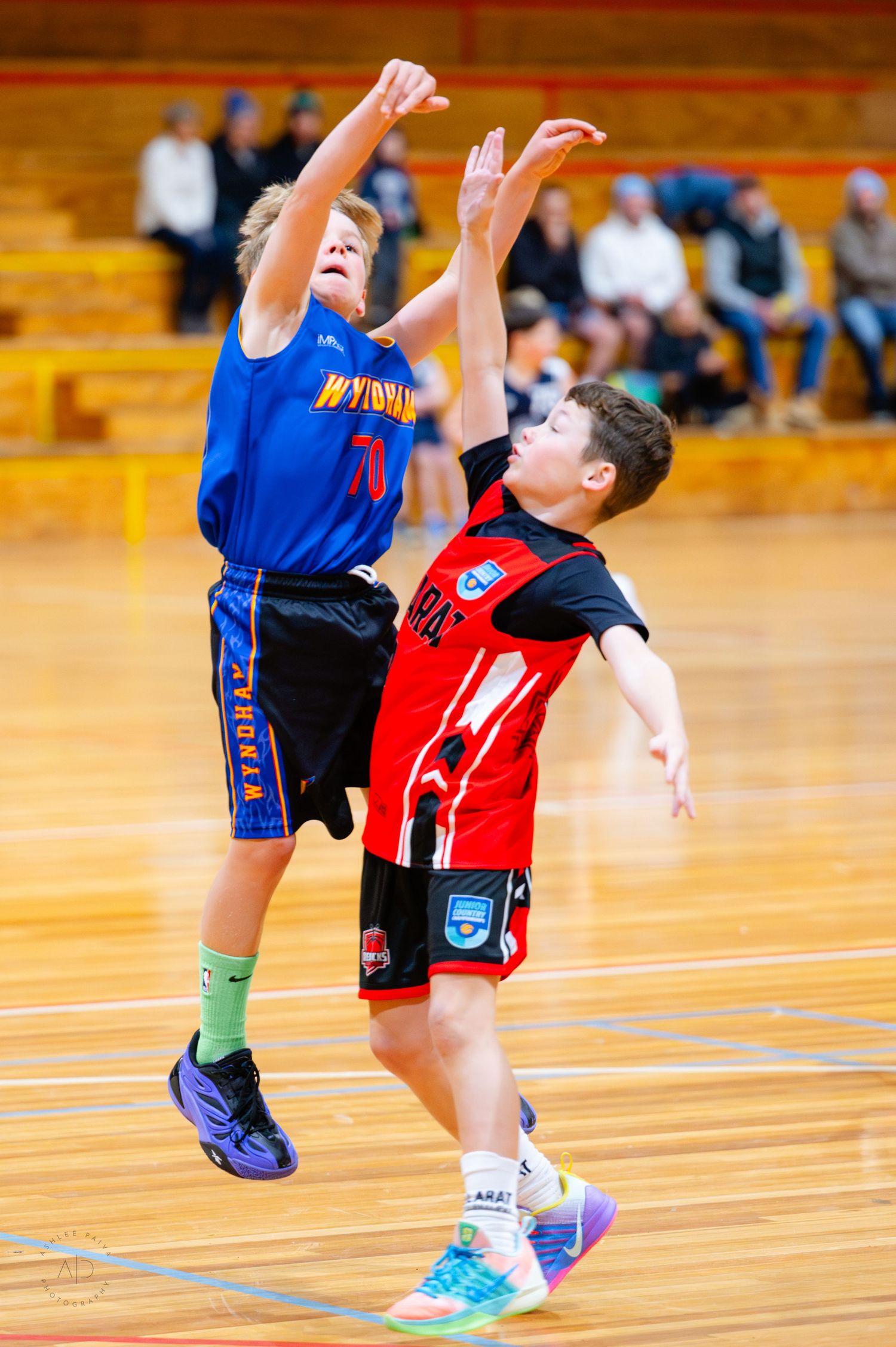 Two young boys playing basketball, one in blue jersey jumping to shoot while opponent in red defends on indoor court.