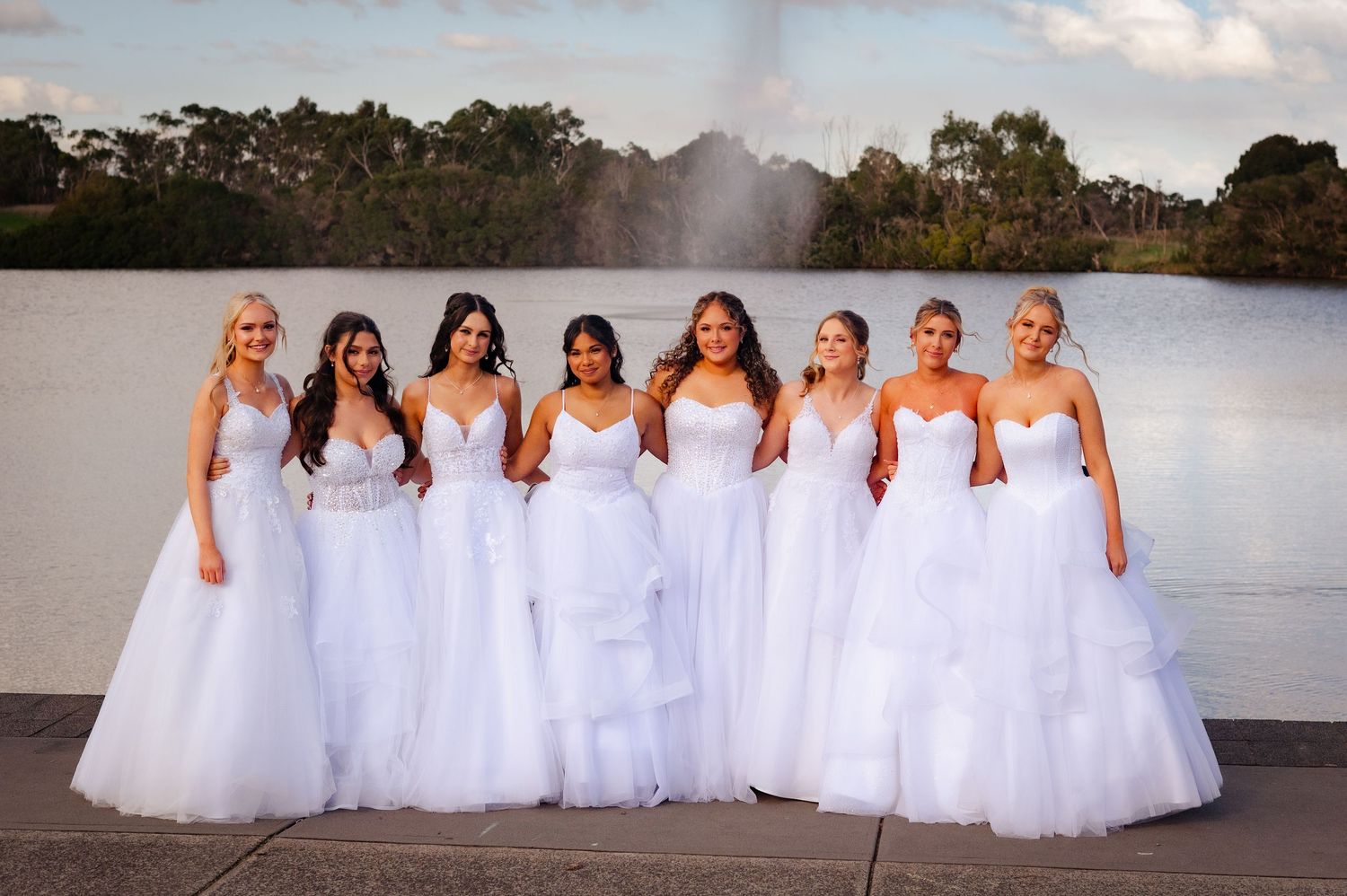 Seven women in elegant white ball gowns pose together outdoors near a fountain, showcasing stunning bridal and formal wear.