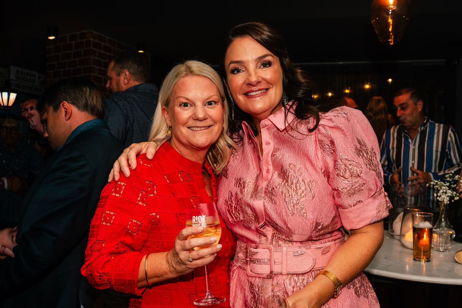 Two women smiling and posing together at a social event, one in red and one in pink dress.