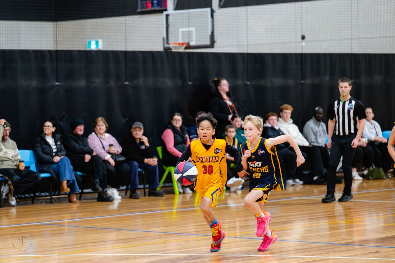 Young basketball players compete in an indoor court game while spectators and a referee watch from the sidelines.