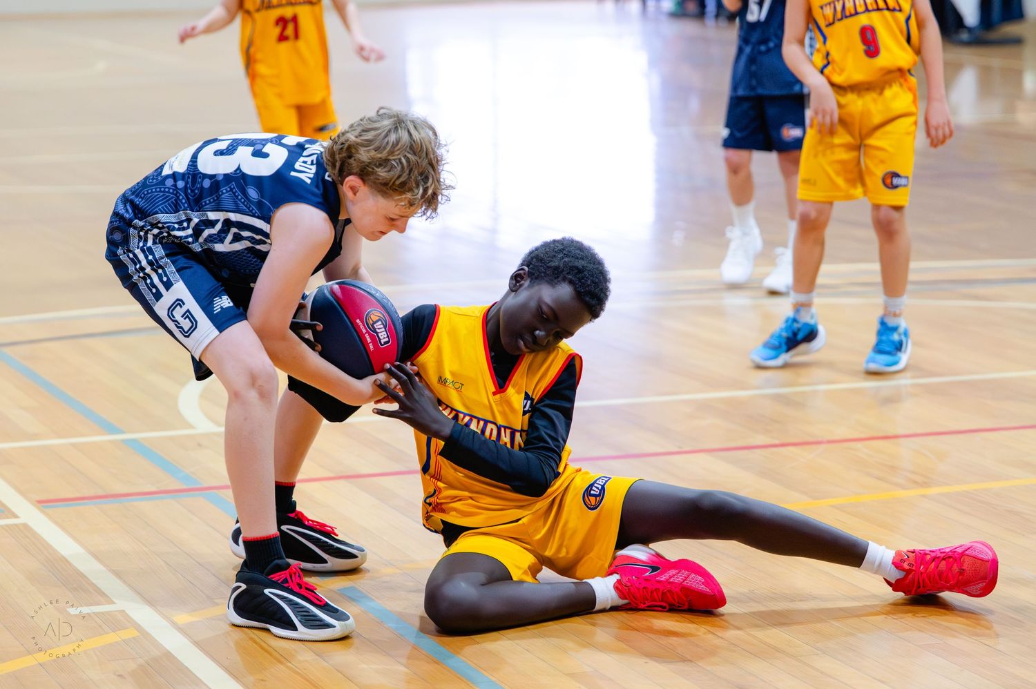 Two youth basketball players compete for the ball during an indoor game, one in blue and one in yellow uniform.