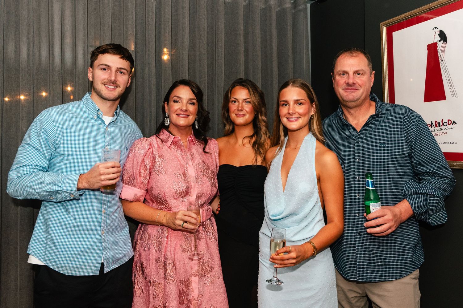 Group of five adults smiling and holding drinks at a social event near a promotional poster.