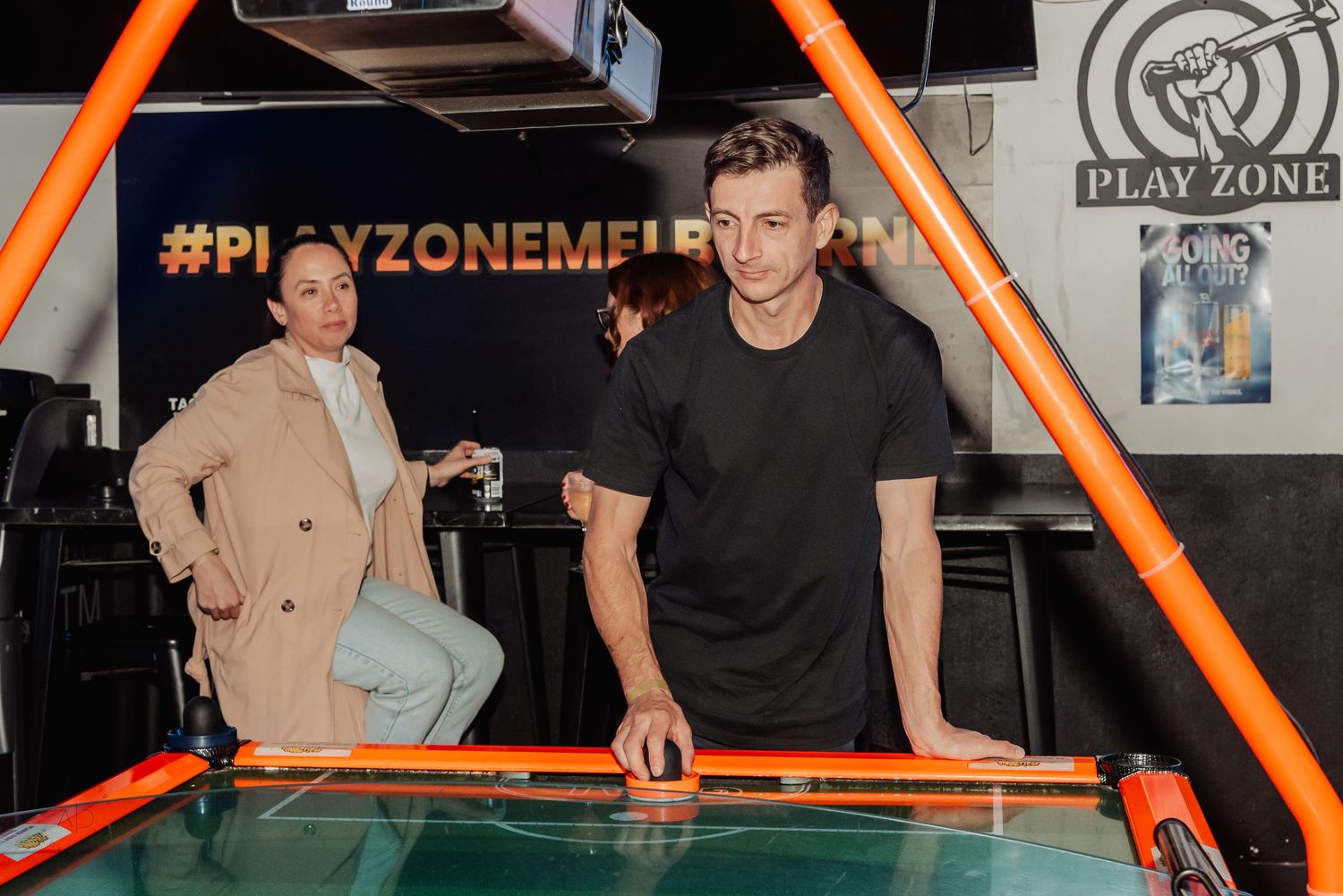 Two people playing air hockey at Play Zone arcade, with orange table and neon lighting in background.