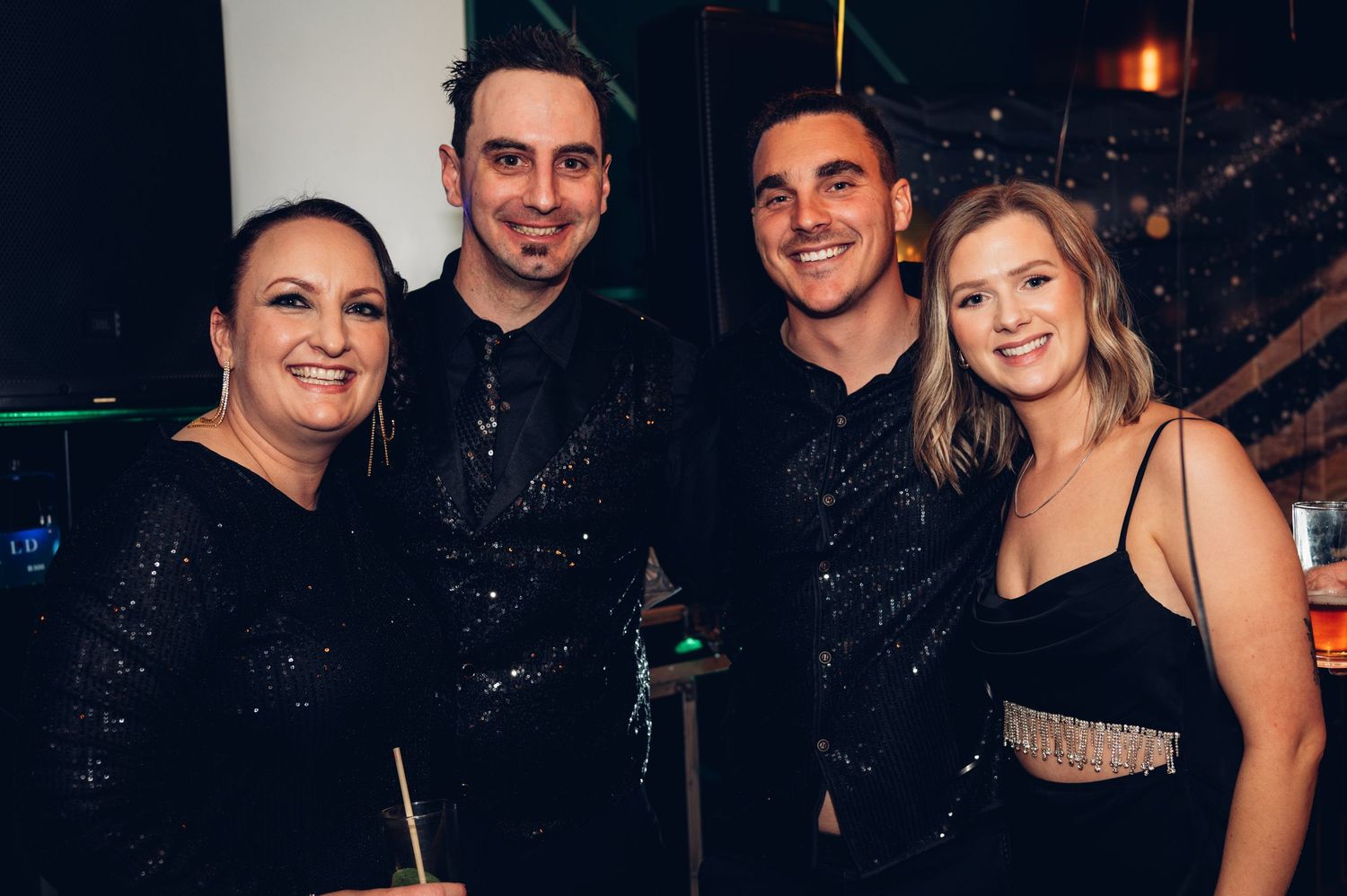 Group of four friends dressed in black attire pose together at a nighttime celebration.