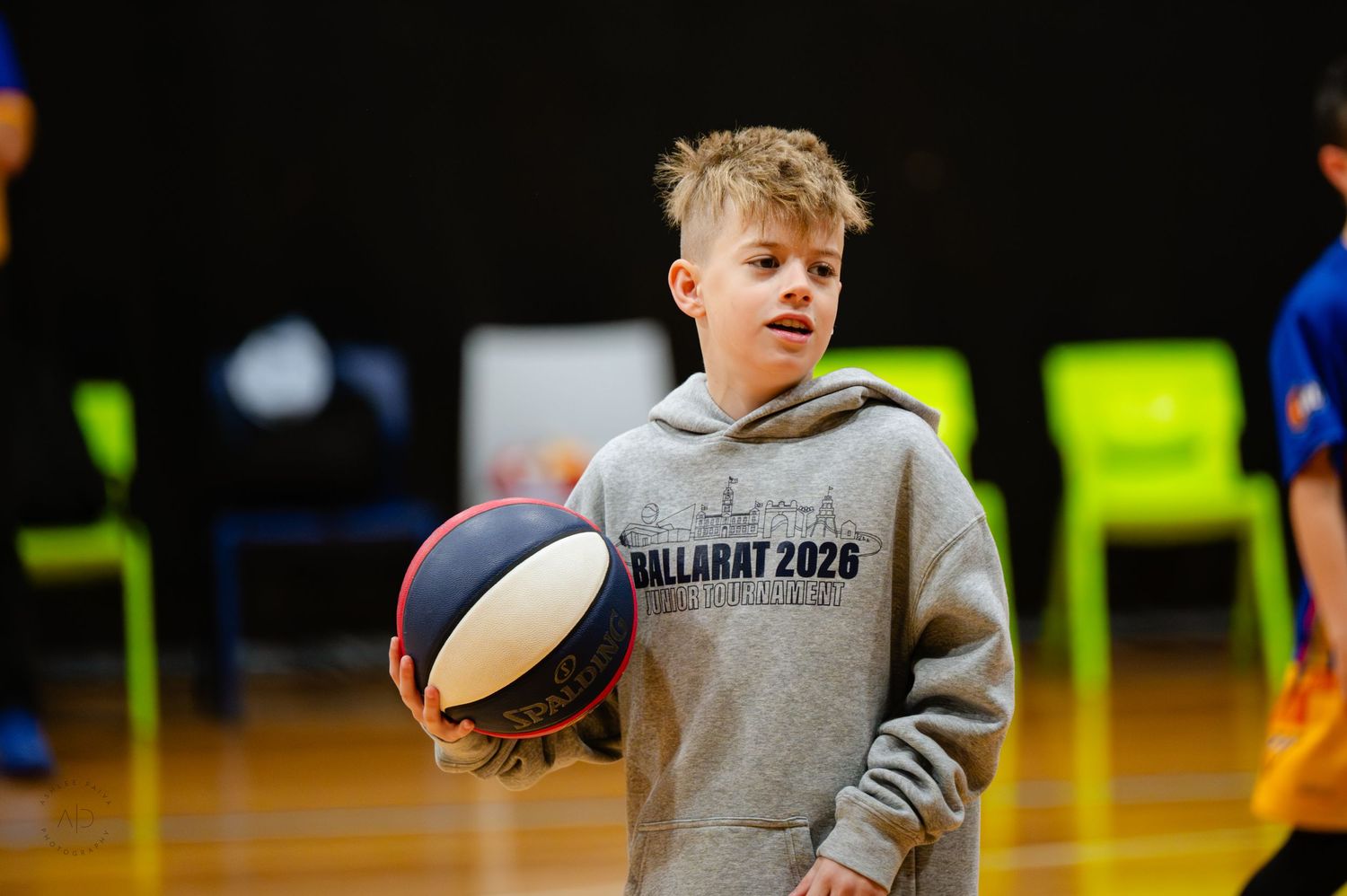Young boy holding a basketball on an indoor court wearing a Ballarat 2026 hoodie.
