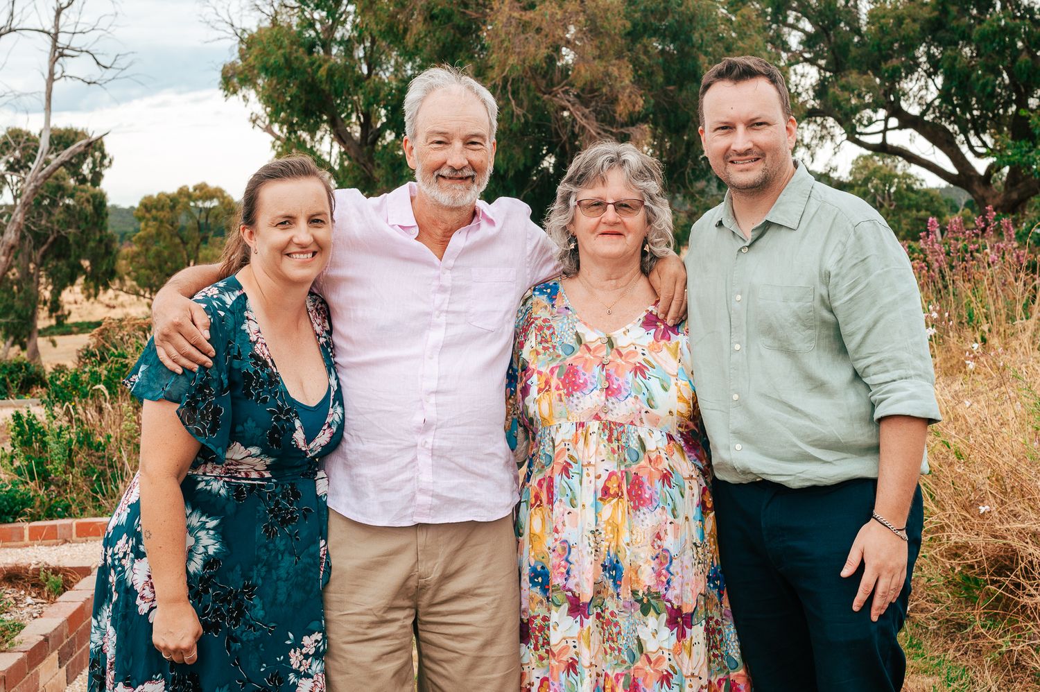 A group of four people stand together outdoors in casual attire against a natural backdrop with trees and tall grass.
