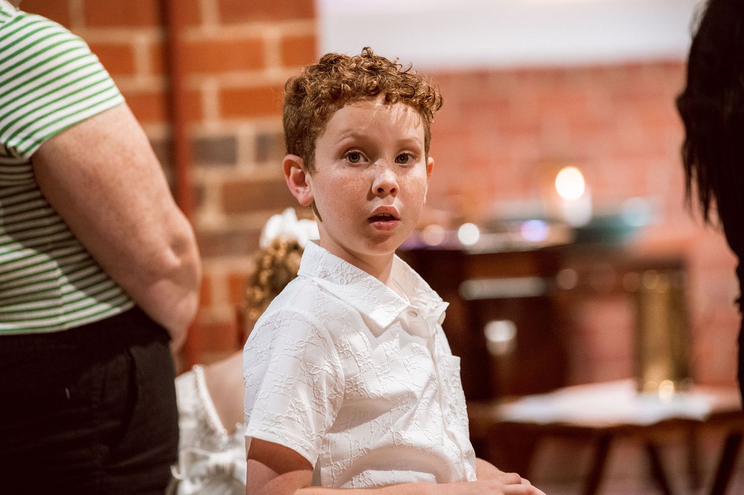 Young child in white clothing stands in a church with brick walls.