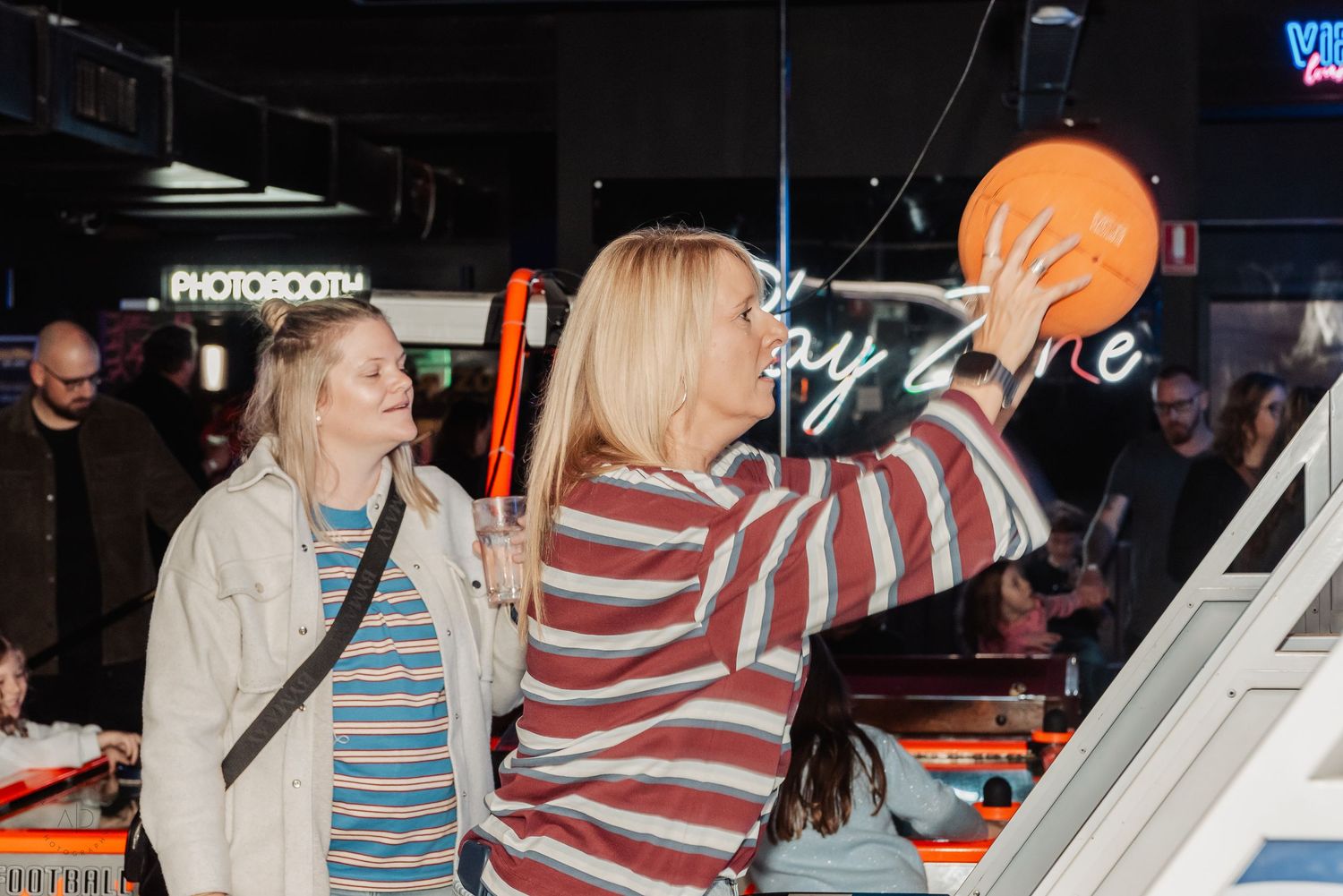 Two women playing skee-ball at a fun entertainment venue with a photobooth and neon signs in background.