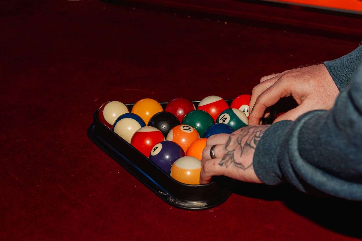 Tattooed hands arranging colorful billiard balls in a rack on a red pool table.