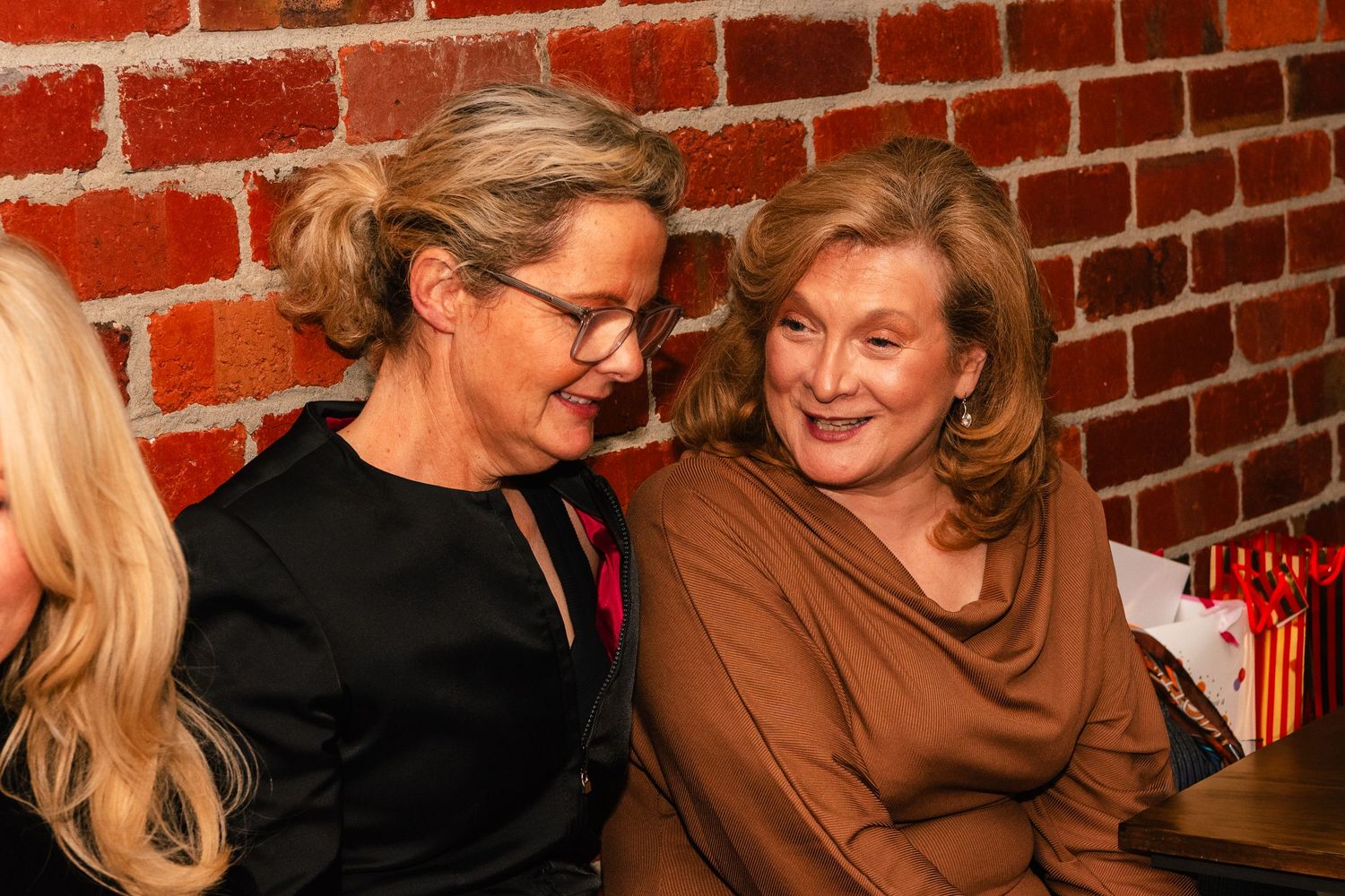 Two elegantly dressed women smiling and chatting together at a social event near a brick wall.